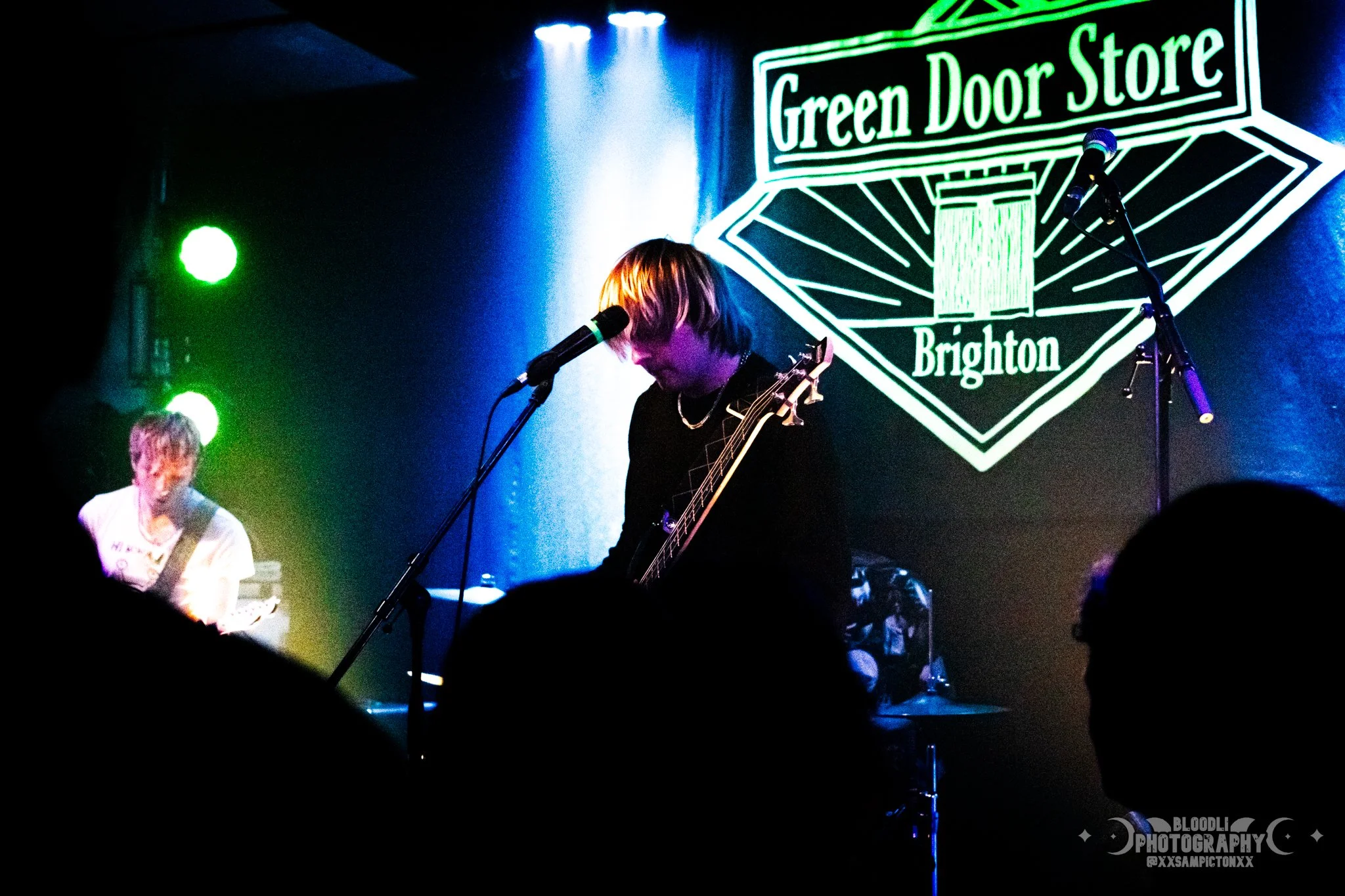 Musicians performing on stage, with one playing guitar and singing into a microphone, at the Green Door Store in Brighton, illuminated by colorful stage lights.