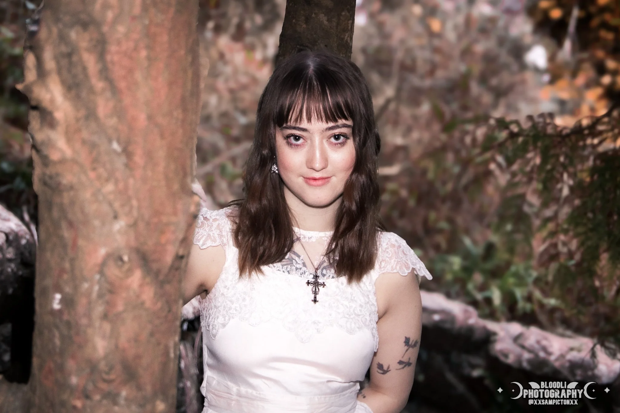 Young woman with wavy brown hair, wearing a white lace dress with lace shoulder details, standing outdoors near a tree, with a background of rocks and foliage, wearing earrings and a cross necklace.