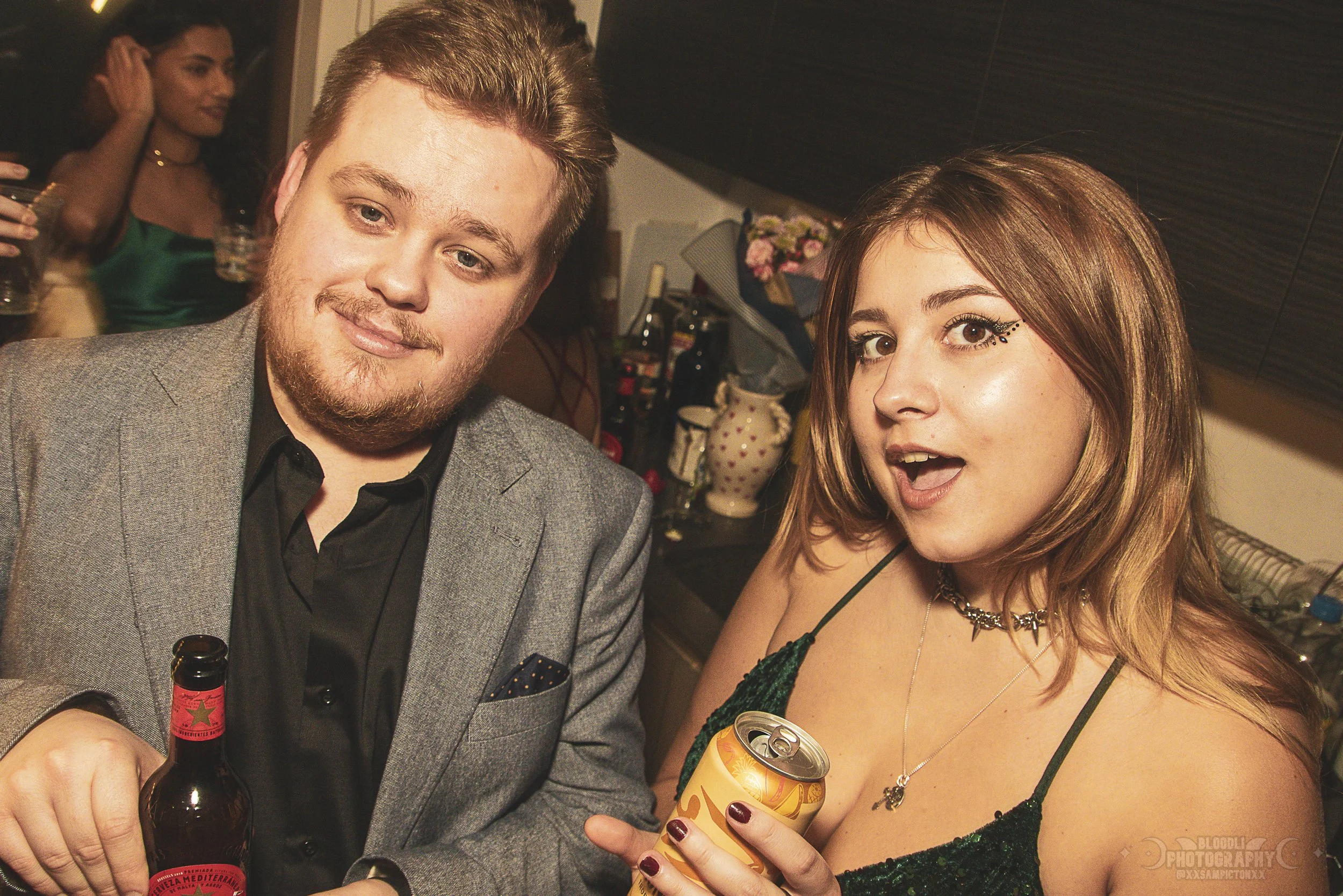 Two young adults at a party, the man in a grey blazer holding a beer and the woman in a green dress holding a can of drink, smiling and posing for the camera.