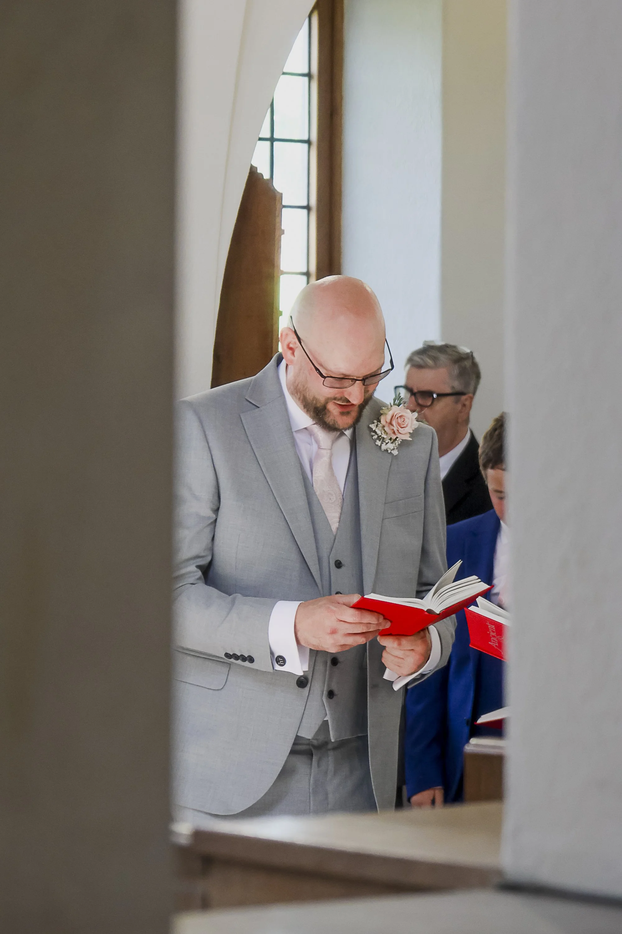 A man with glasses, wearing a gray suit and a boutonniere, is reading from a red book during a wedding ceremony, standing beside a boy in a blue suit, with people and a window in the background.