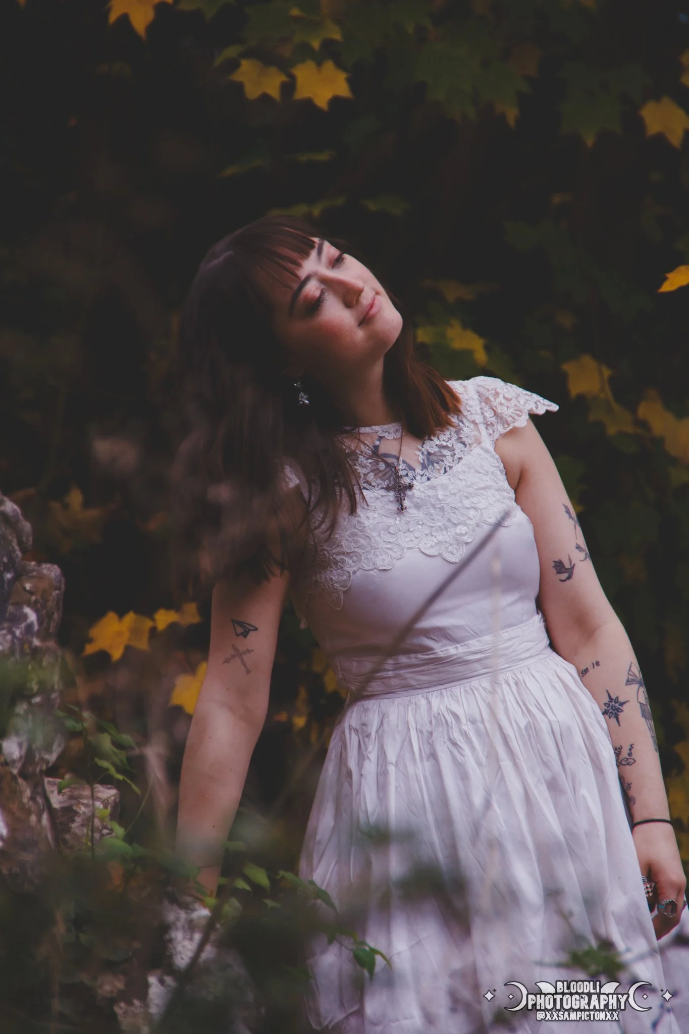 A young woman in a white dress with lace details stands outdoors among green and yellow leaves, with her eyes closed and head tilted slightly to the side, in a peaceful pose.