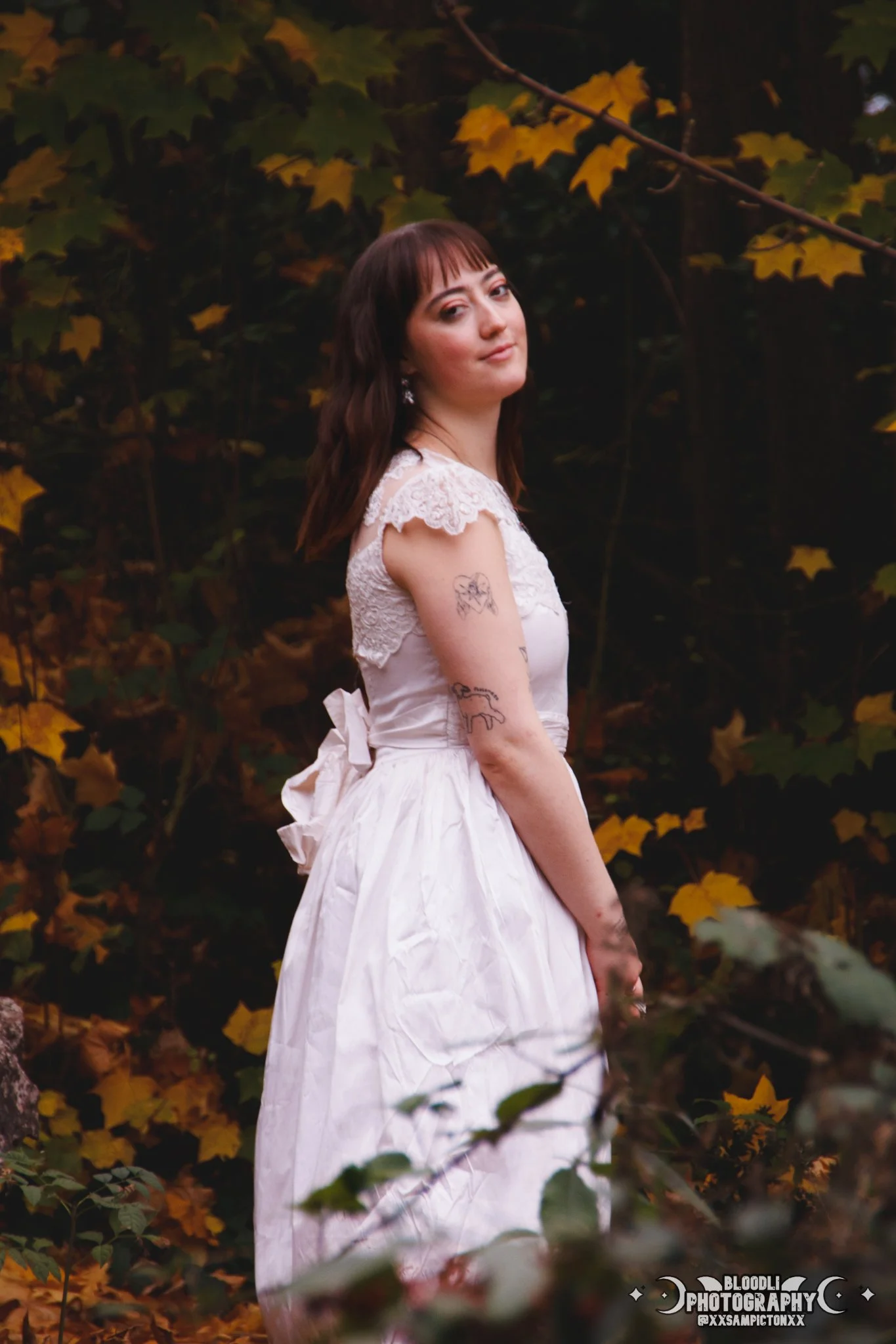 A young woman in a white dress with lace details and a bow on the back standing in a forest with autumn leaves. She has dark brown hair, tattoos on her left arm, and is looking at the camera with a slight smile.