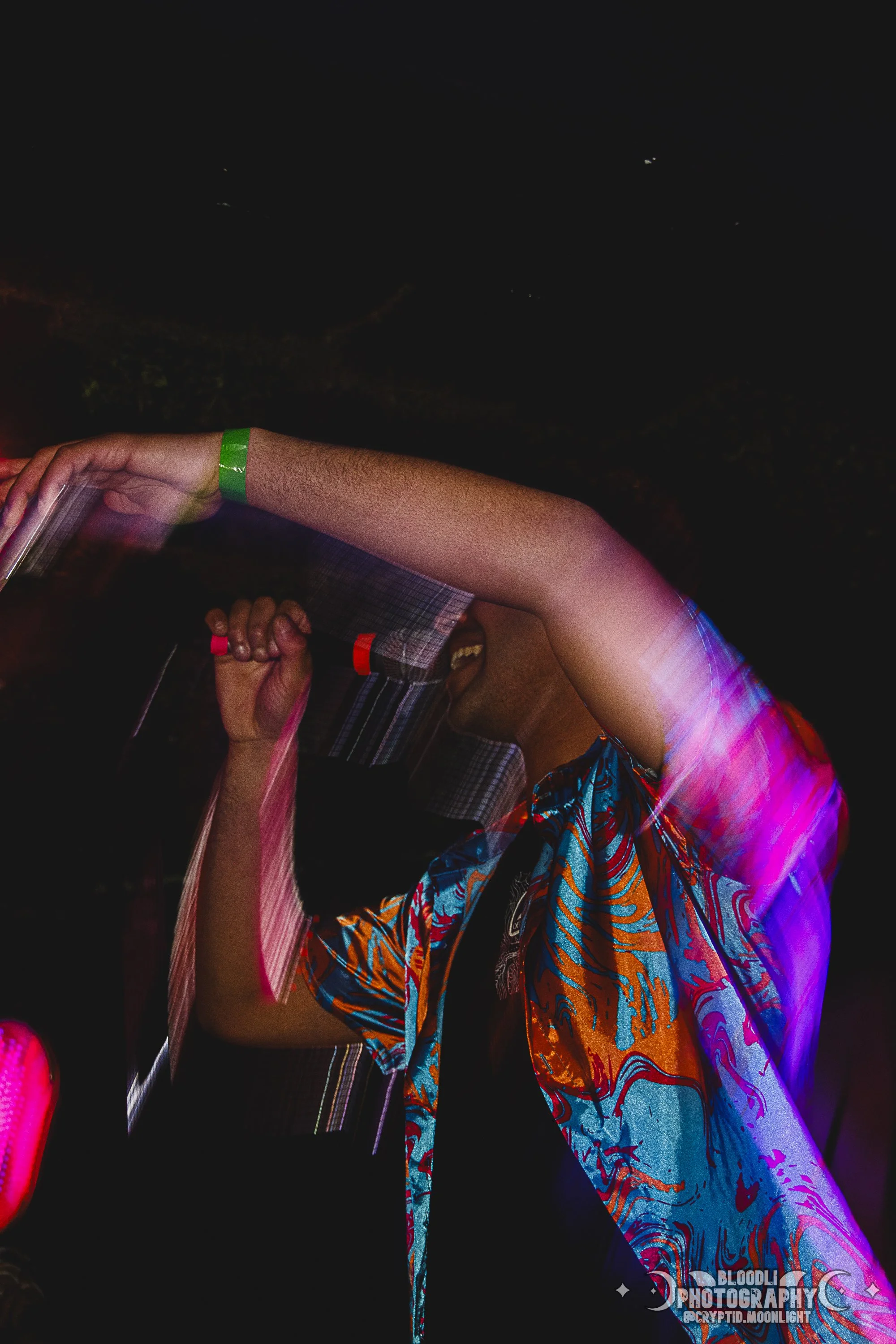 A person dancing at a party or club, wearing a colorful, patterned shirt and a green wristband, with a dark background and light streaks.