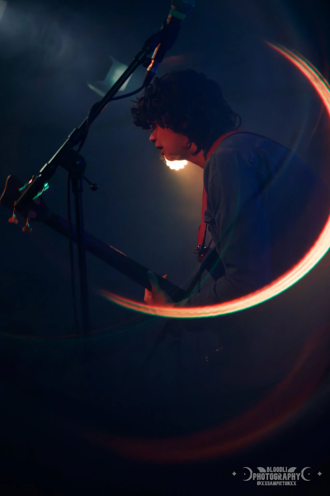 A person with curly hair playing an acoustic guitar on stage at night, illuminated by a bright stage light.