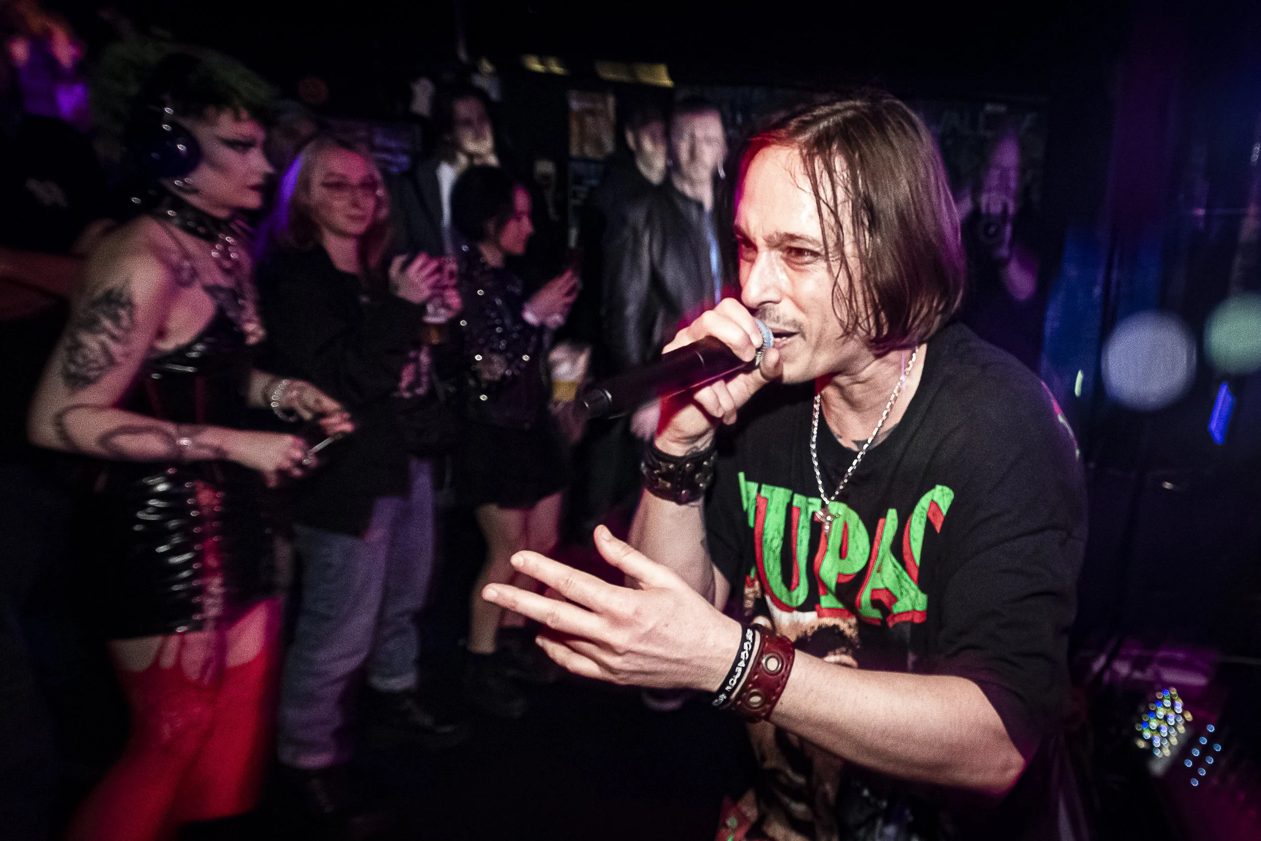 A male performer with shoulder-length brown hair singing into a microphone at a concert, wearing a black band T-shirt with green and red lettering, and surrounded by an audience in a dark venue.