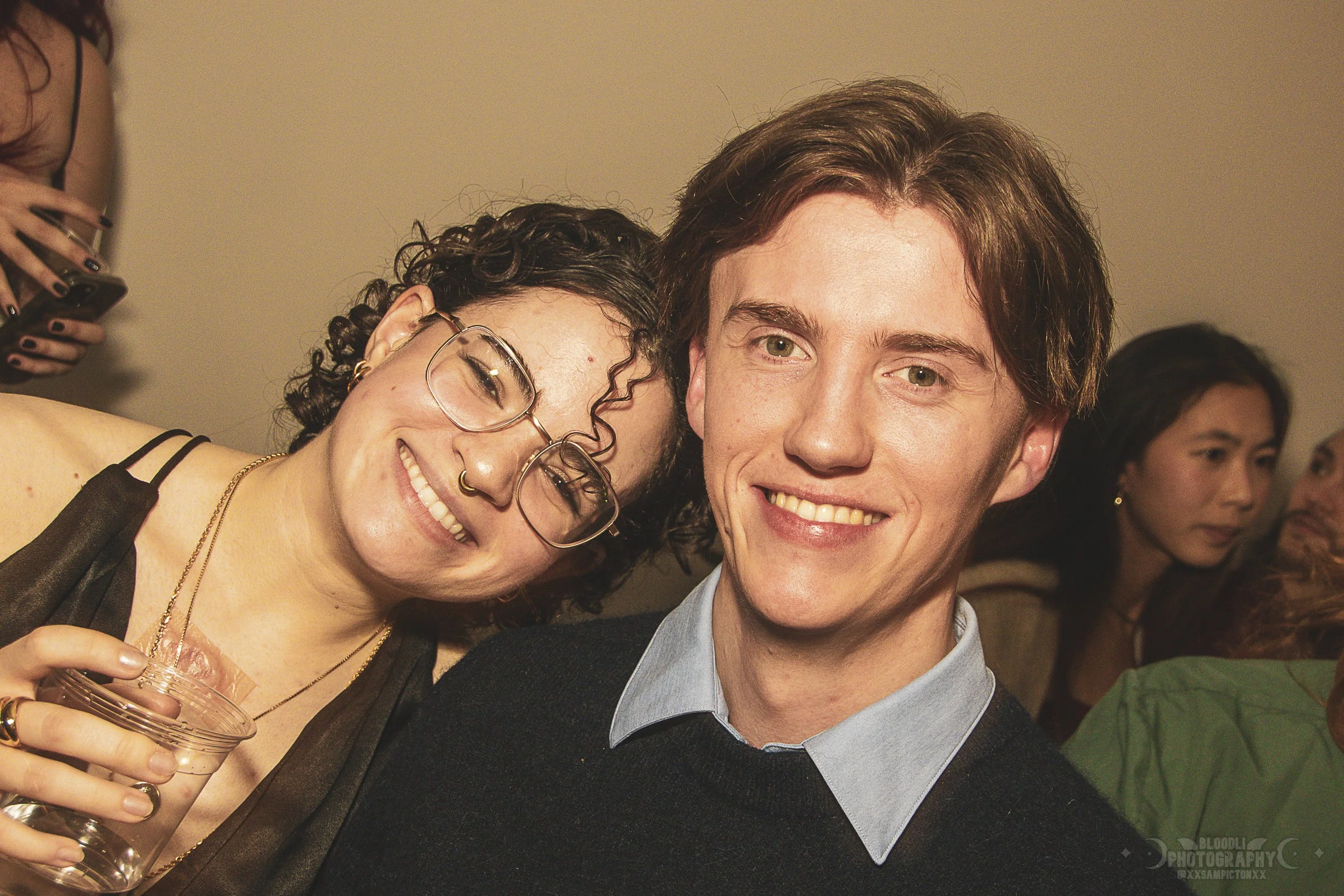 A young man and woman smiling at a social gathering, with the woman wearing glasses, a nose ring, and holding a drink, and people in the background