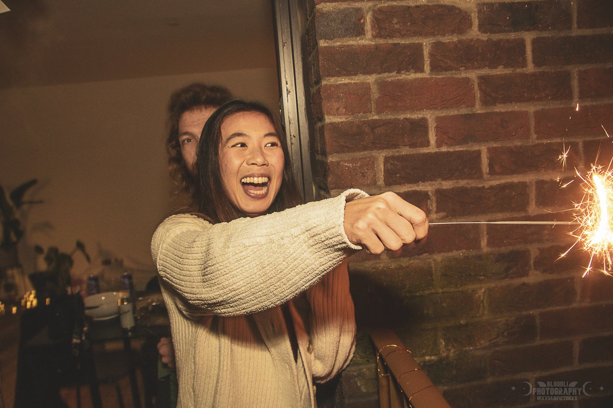 A woman and a man celebrating indoors, with the woman holding a sparkler near a brick wall, smiling and enjoying the moment.