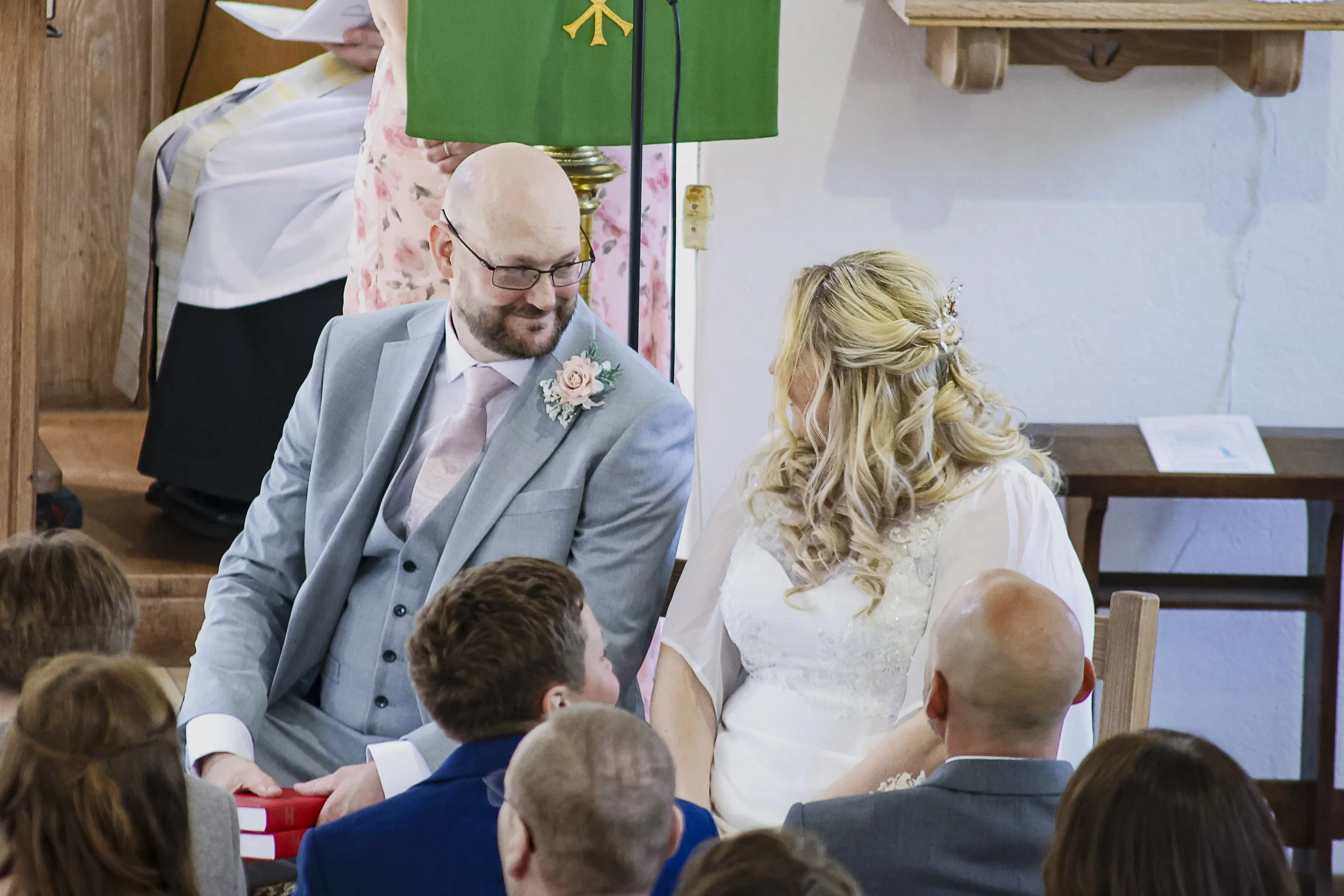 A wedding ceremony with a bride and groom sitting at a table, surrounded by guests in a church setting.