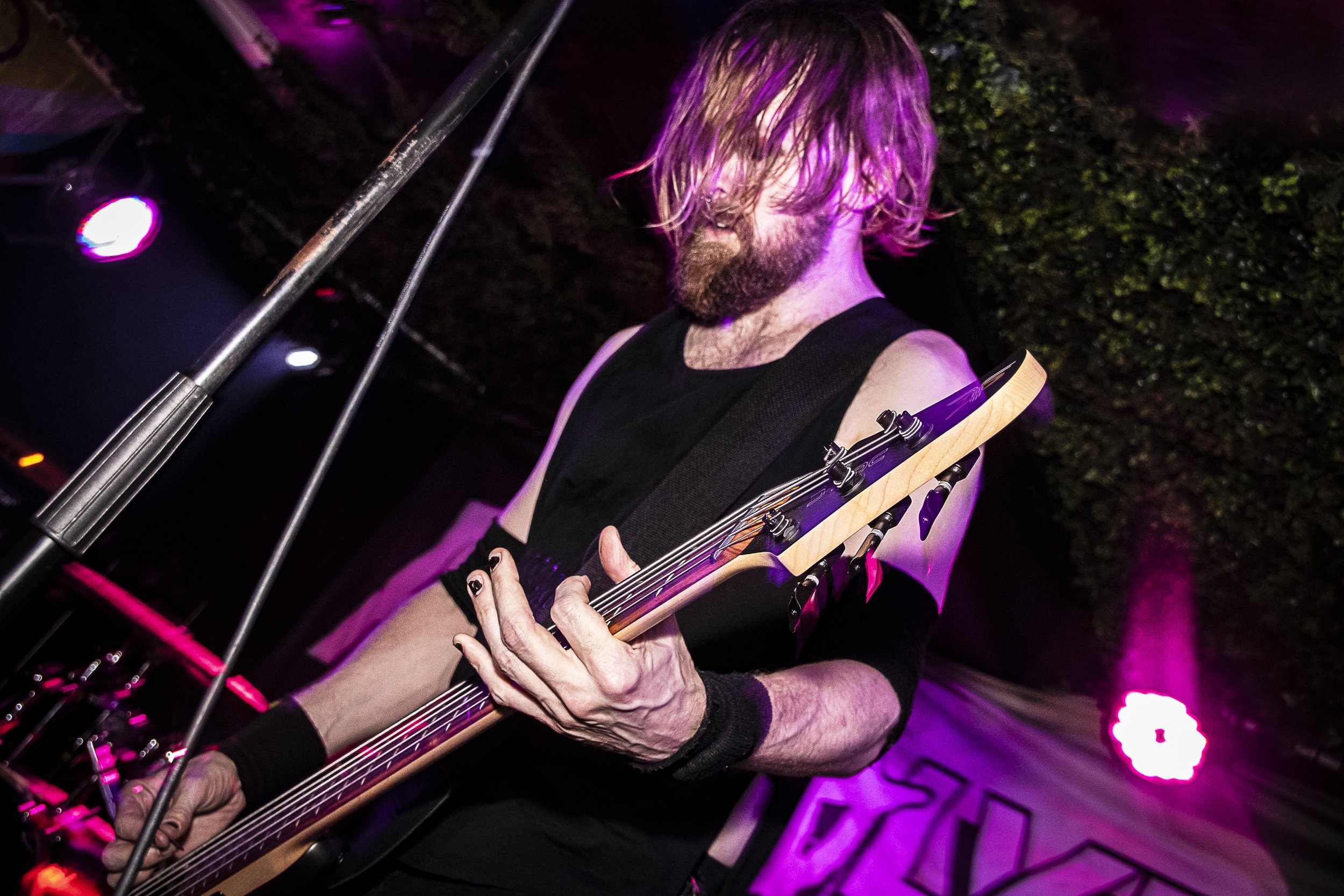 A bearded man with long hair, wearing a black sleeveless shirt, playing an electric bass guitar on stage with purple lighting.