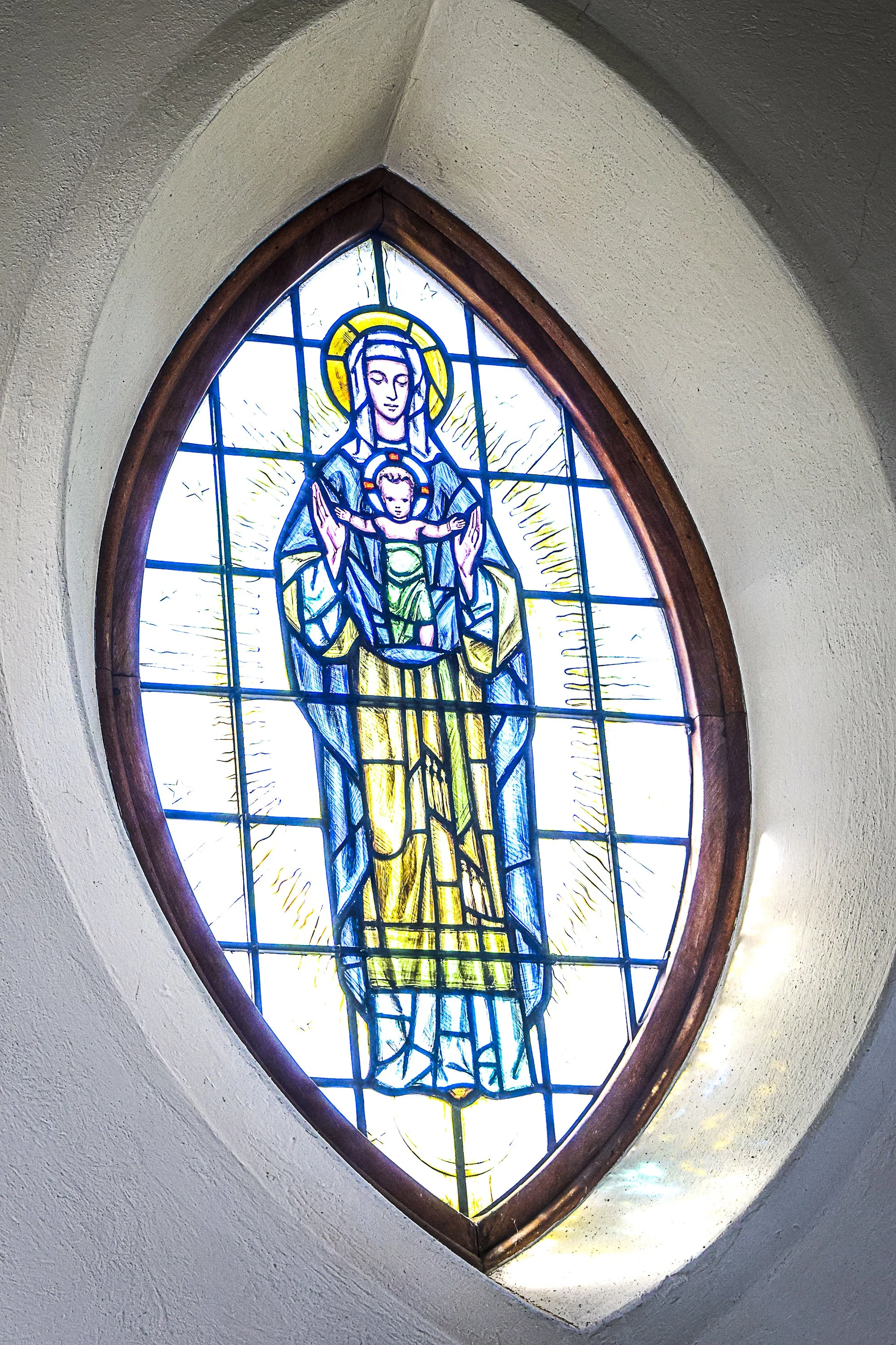 Stained glass depiction of the Virgin Mary holding Baby Jesus, with a bright yellow halo, in a church window.