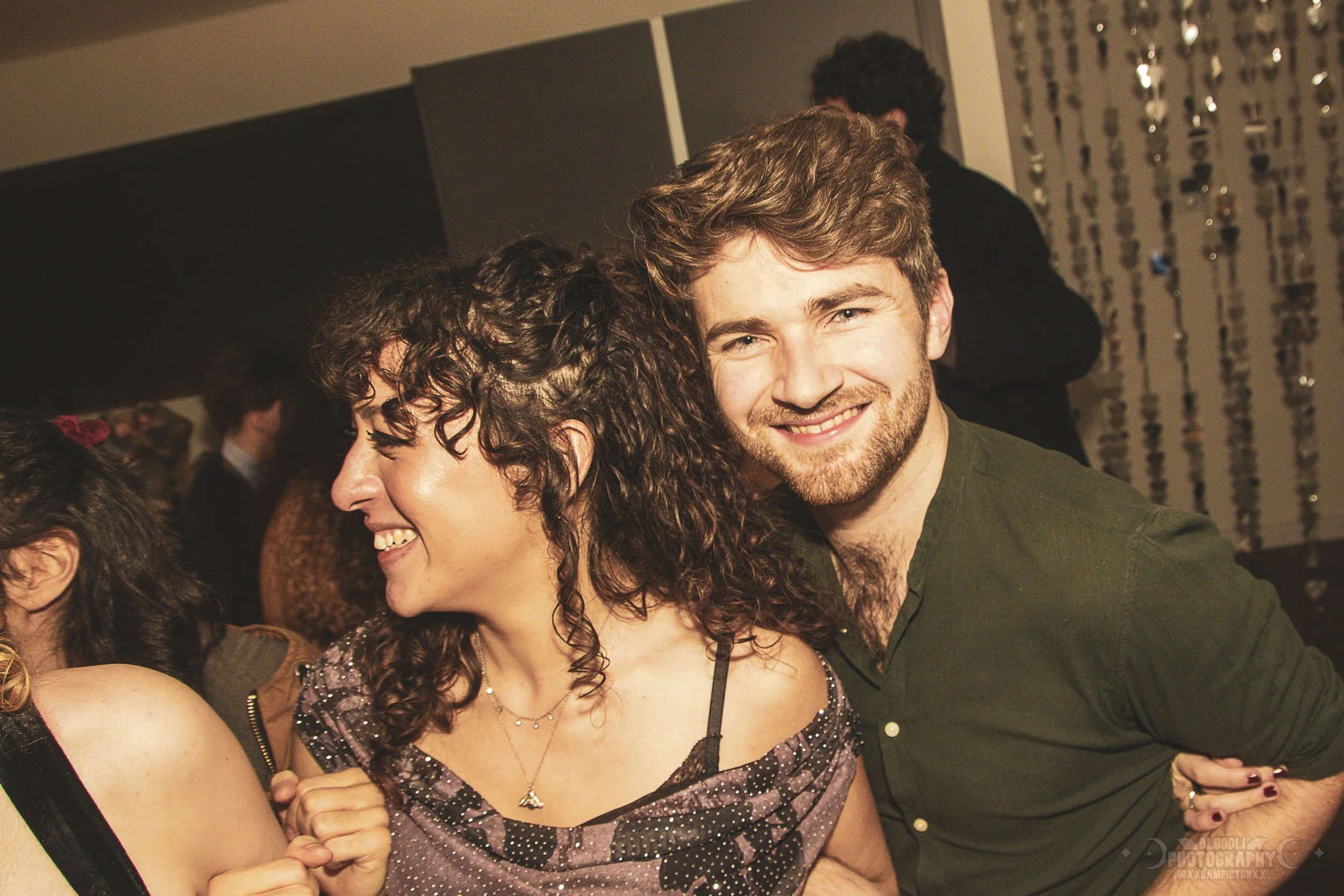 Two young adults, a woman with curly dark hair and a man with light brown hair and a beard, smiling and embracing at a party or social gathering.