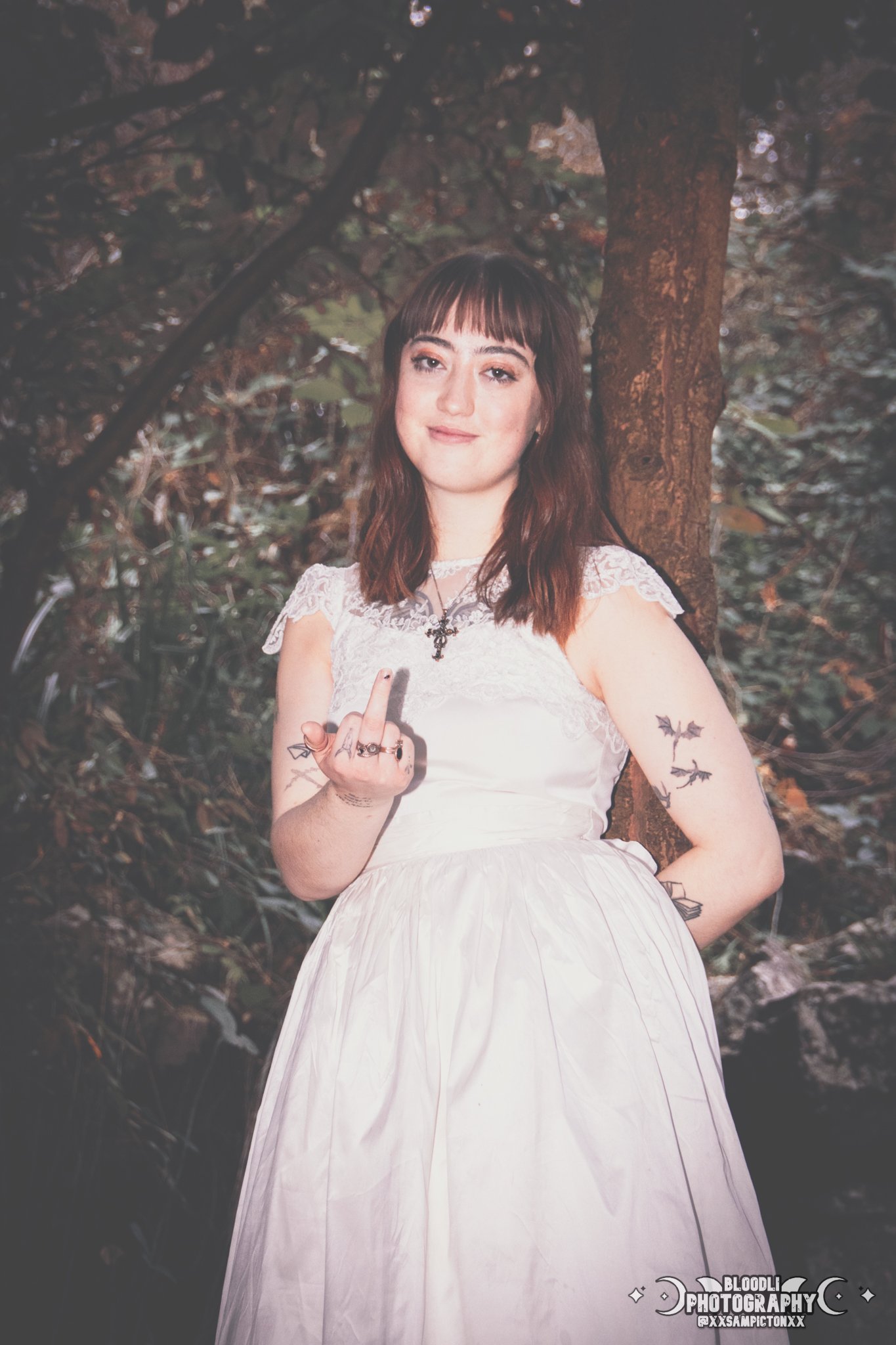 A young woman with dark brown hair and bangs, wearing a white dress with lace details, stands outdoors in front of a tree with dark leaves. She has tattoos of bats on her arms and is holding up her middle finger toward the camera, with a subtle smile