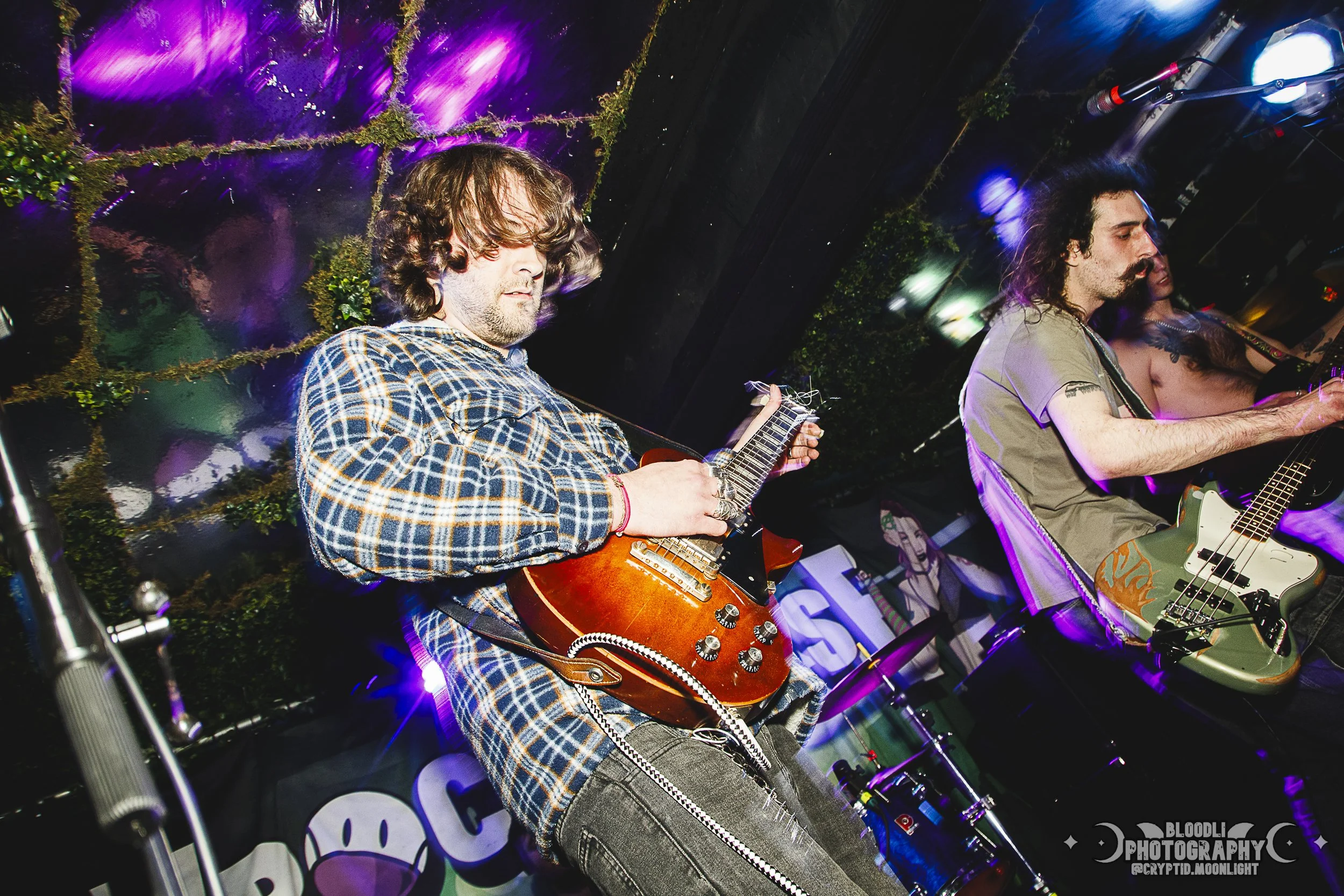 Two male musicians playing guitars on a stage with colorful lights and a black background. One is wearing a plaid shirt, and the other a casual t-shirt.