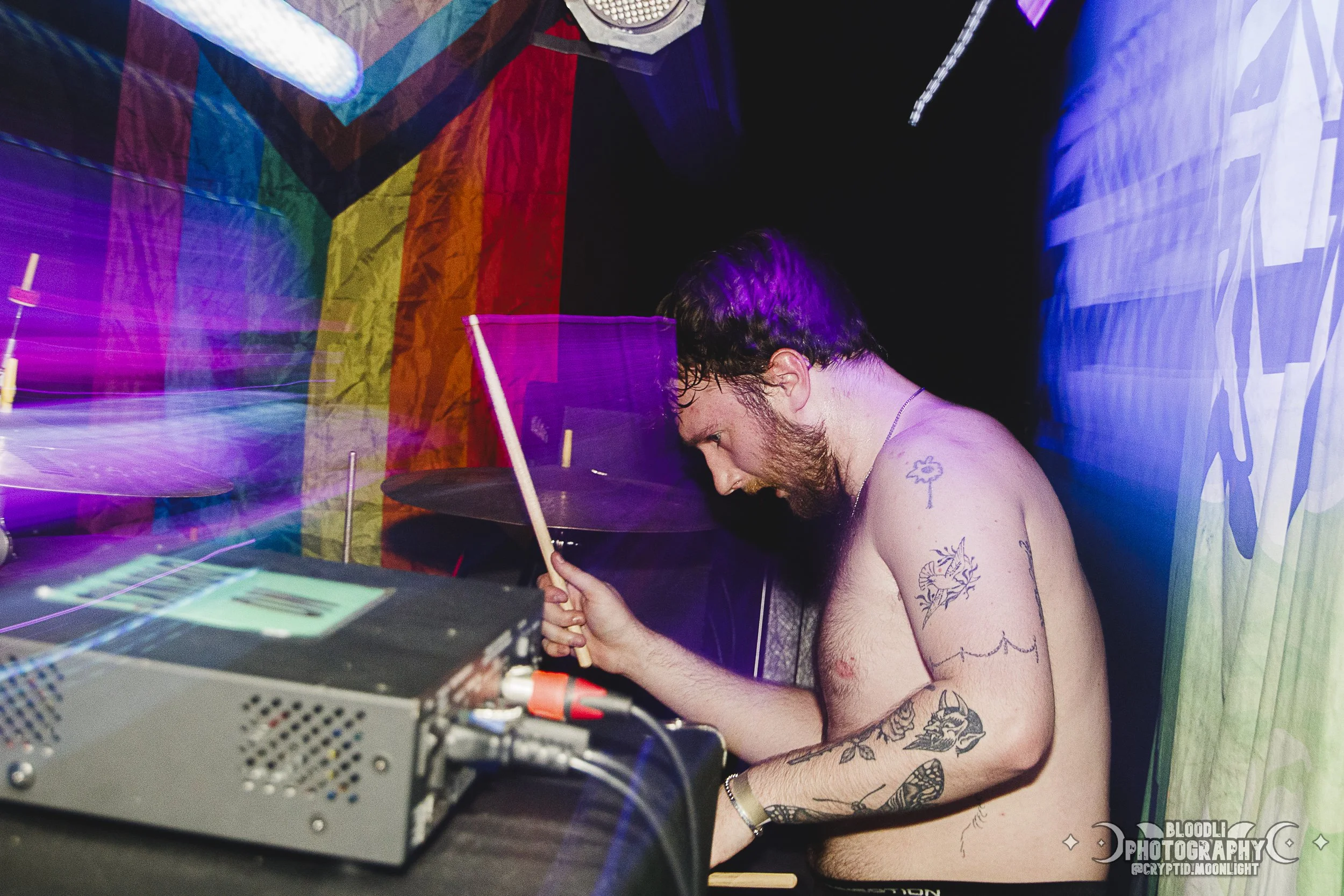 A shirtless male musician with tattoos on his arm, playing drums at a colorful, vibrant indoor concert or party. The background features rainbow-colored flags or banners, and the scene is lit with purple and pink lights, creating a dynamic and energe