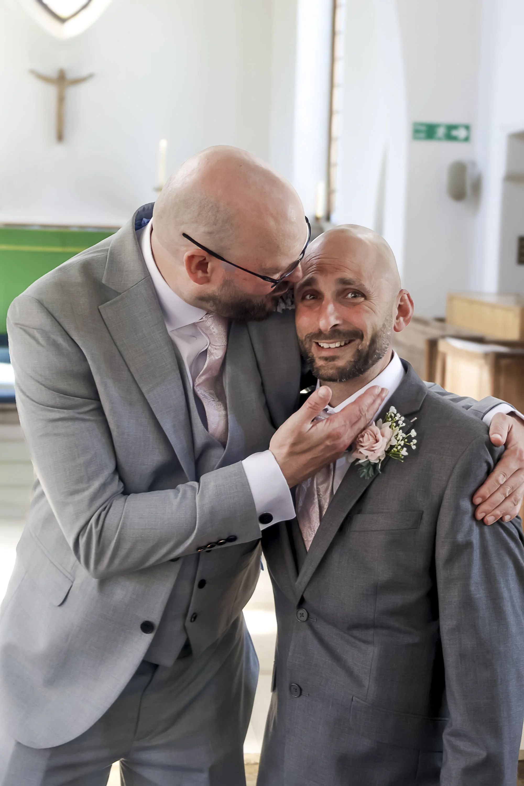 Two men in suits sharing a joyful moment, one kissing the other on the forehead while they smile and hold each other at a formal event, likely a wedding.