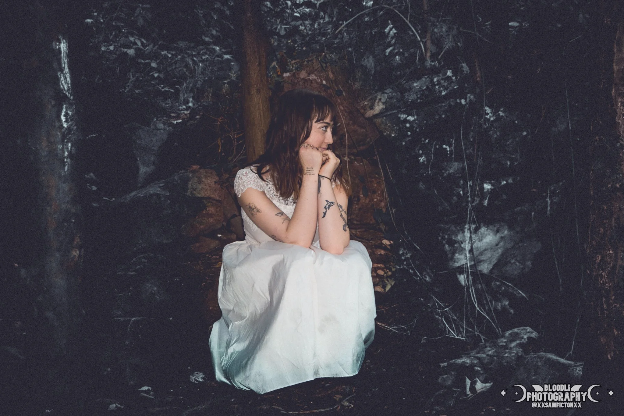 A young woman with tattoos, wearing a white dress, sitting on the ground in a cave or rocky environment, with her chin resting on her hands, looking to her left.
