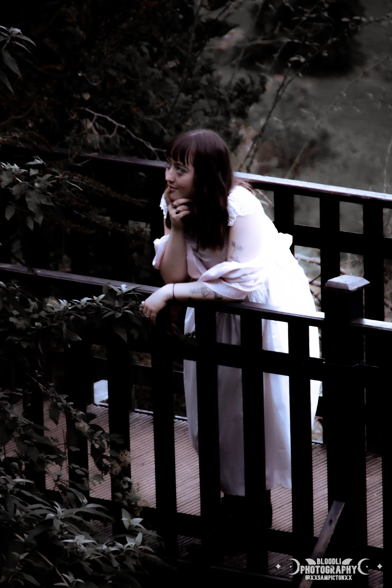 A woman with dark hair wearing a white dress leaning on a wooden railing, looking thoughtfully into the distance in a natural setting.