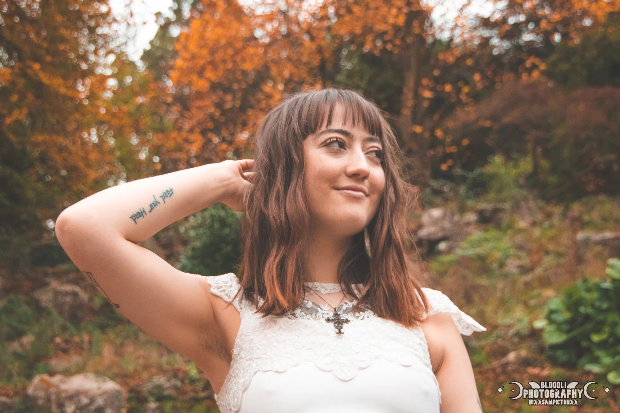 Young woman with shoulder-length brown hair and bangs standing outdoors in autumn with trees and rocks in the background, wearing a white lace top and a cross necklace, smiling slightly with her right hand behind her head.