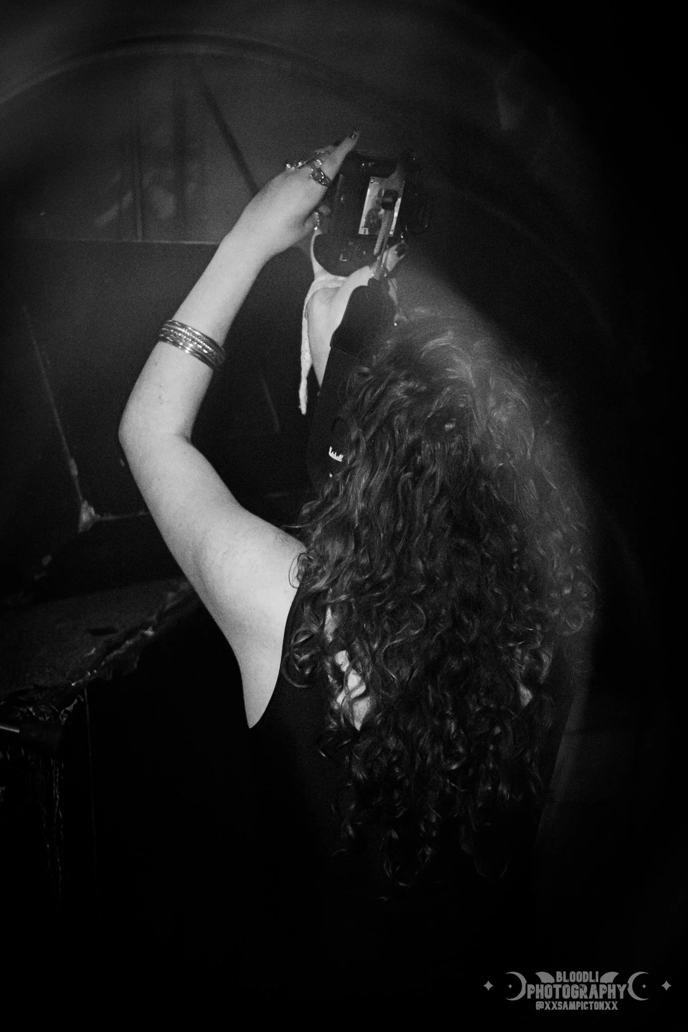 A woman with curly hair taking a photo with a camera inside a dark room, with her back to the camera, wearing bracelets.