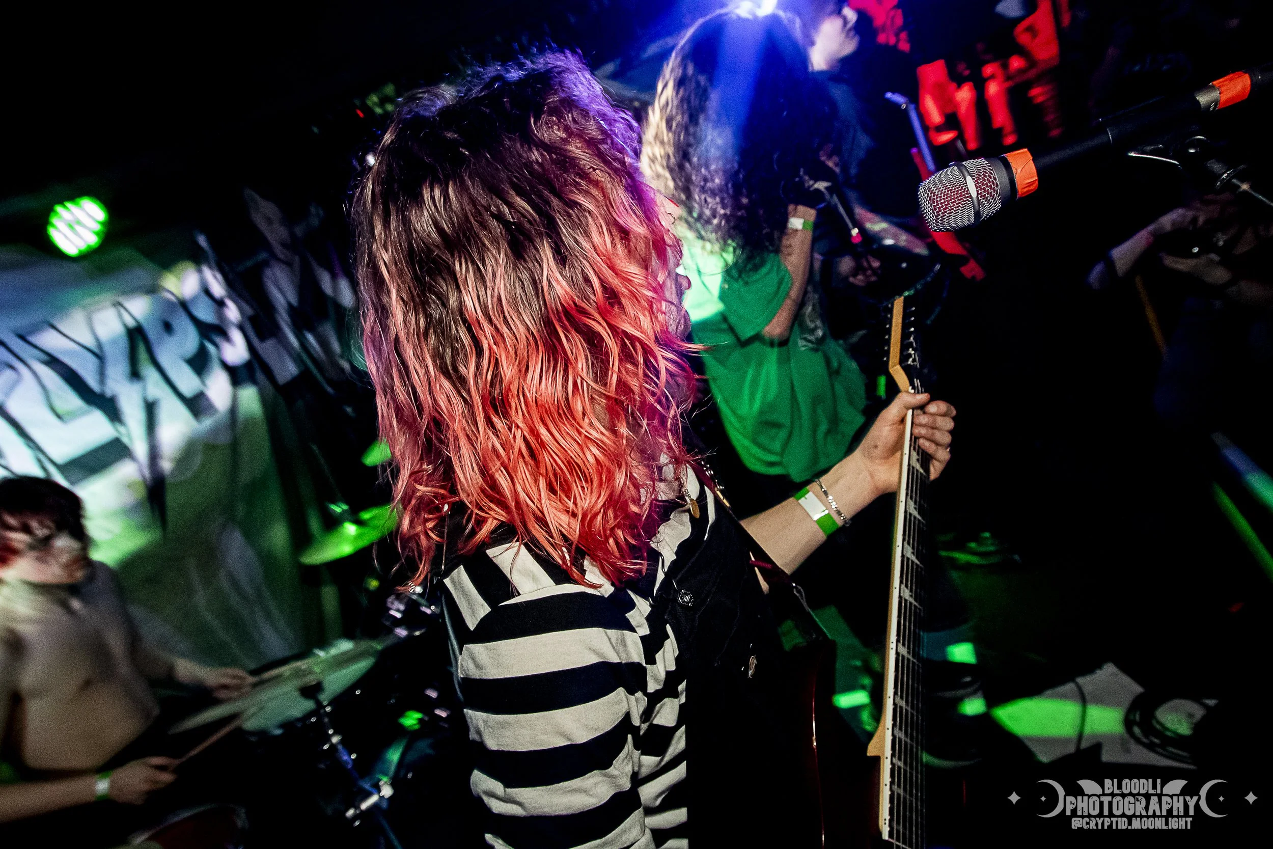 Person with wavy hair dyed pink and purple, wearing a black and white striped shirt, playing guitar on stage with colored lights. Other band members are visible in the background.