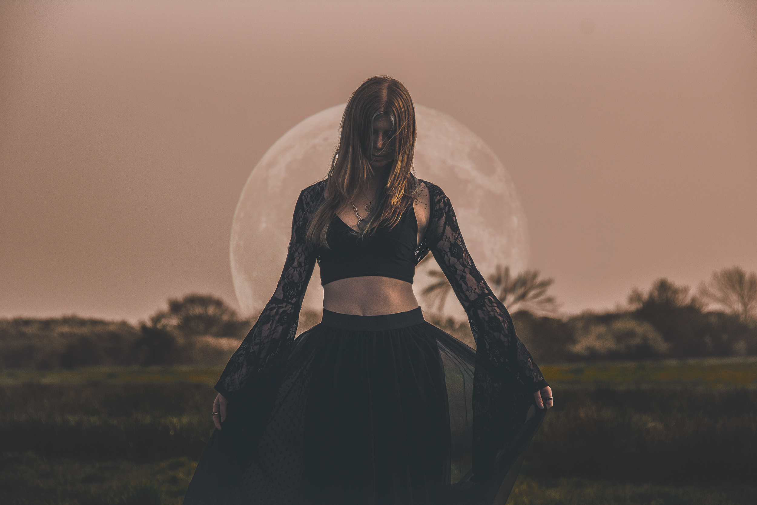 A woman in a black lace outfit standing outdoors at sunset with a large full moon in the background.