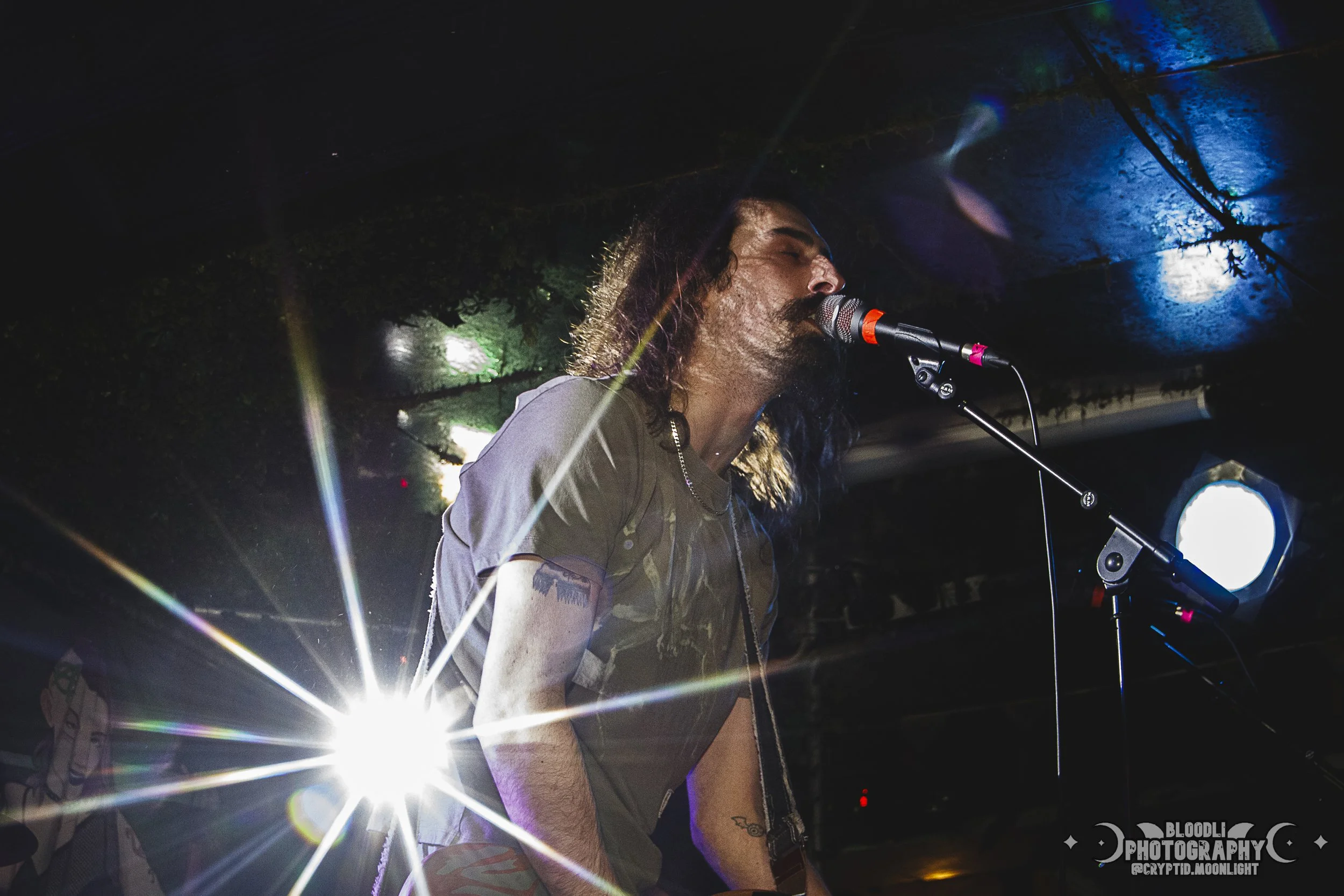 A man with long hair and a beard singing into a microphone on stage, with colorful lights and a bright backlight creating a lens flare.