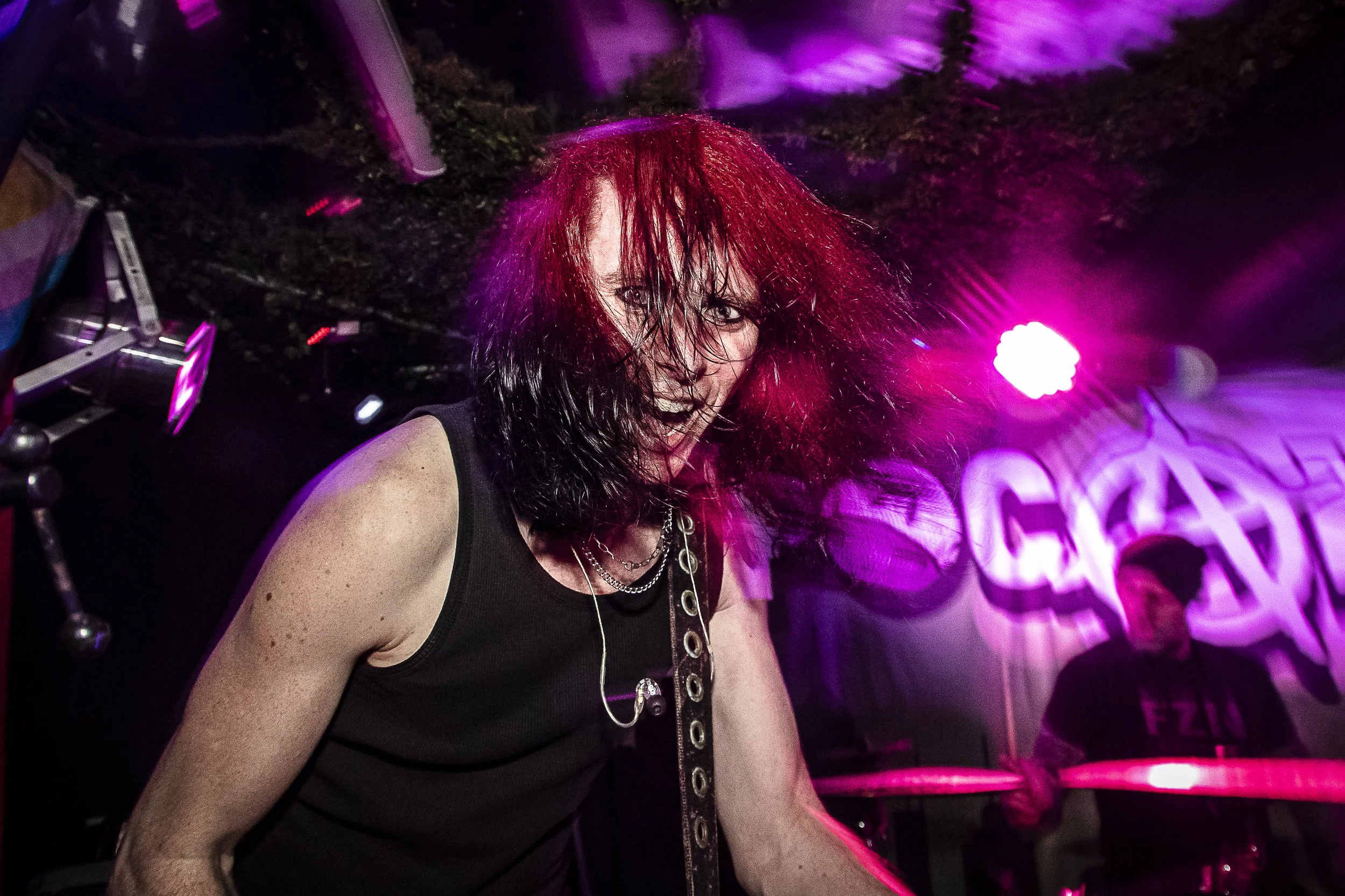 A woman with wet, black and red hair, wearing a sleeveless black top, performing with a guitar at a live music venue with purple stage lighting.