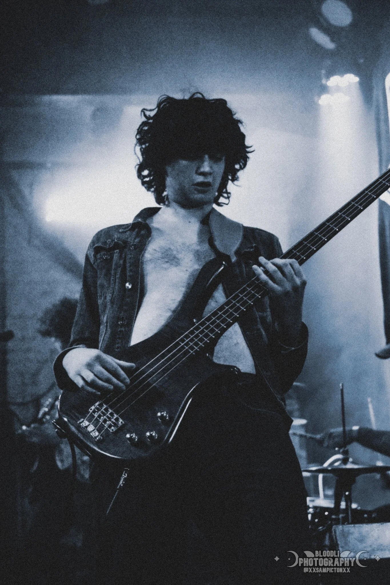 A woman playing an electric bass guitar in a dimly lit room, with curly hair and an open shirt.