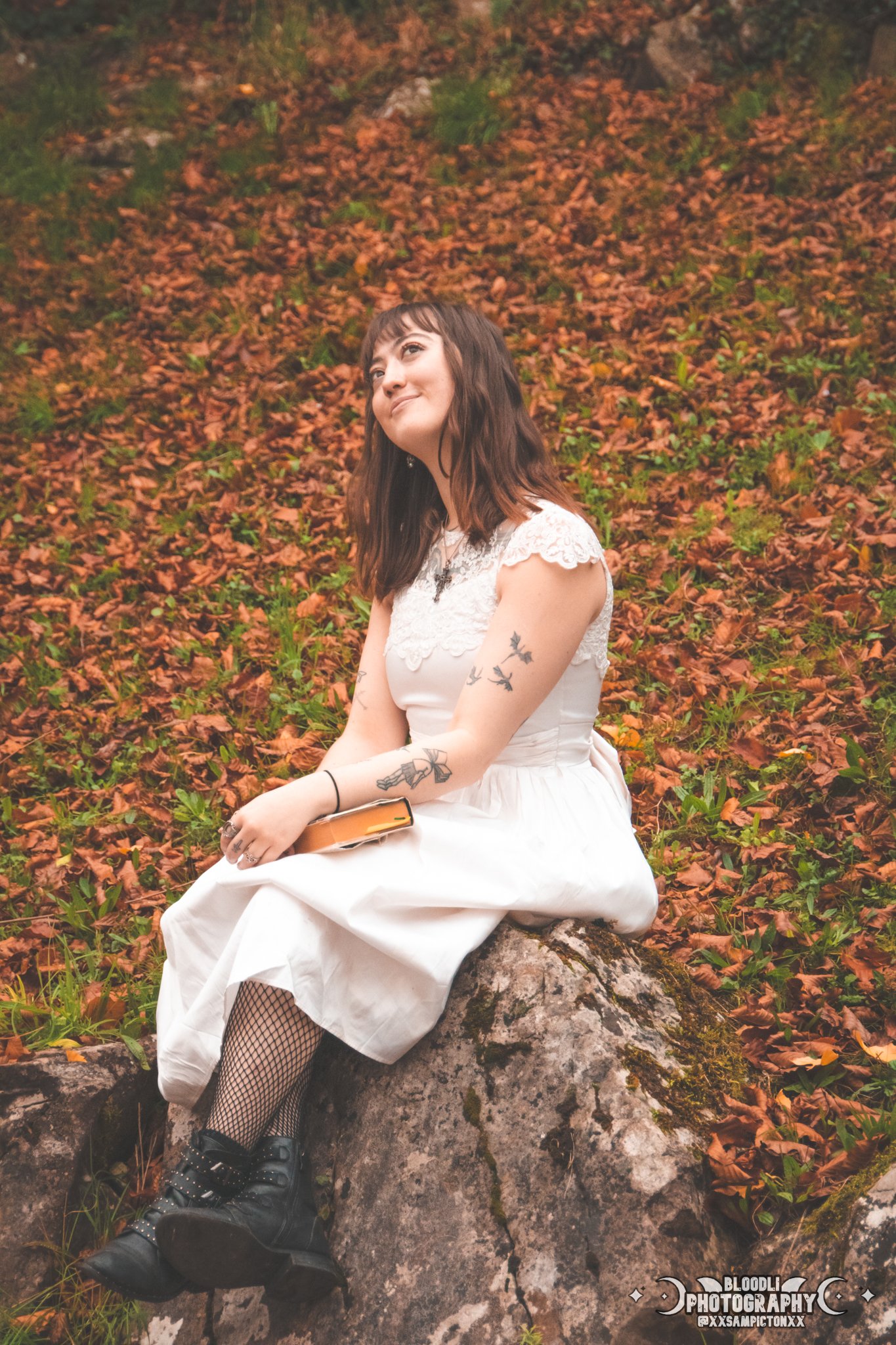 A young woman with dark hair and tattoos, wearing a white dress with lace sleeves, fishnet stockings, and black boots, sitting on a rock amidst autumn leaves in a forest, holding a book and smiling thoughtfully.