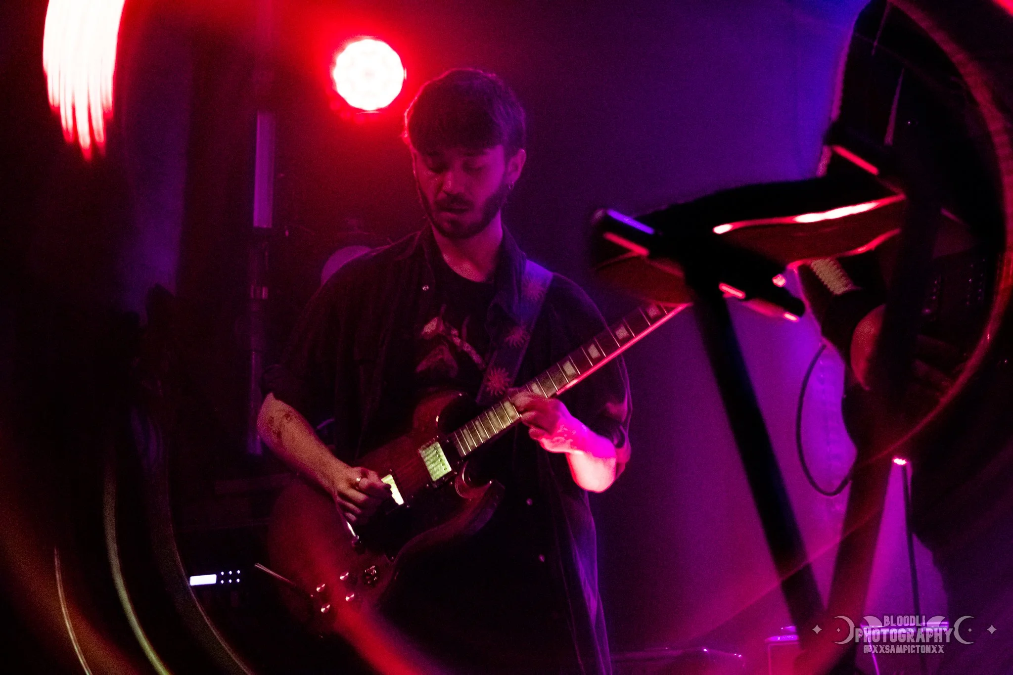 A musician playing an electric guitar on stage under red and purple lighting.
