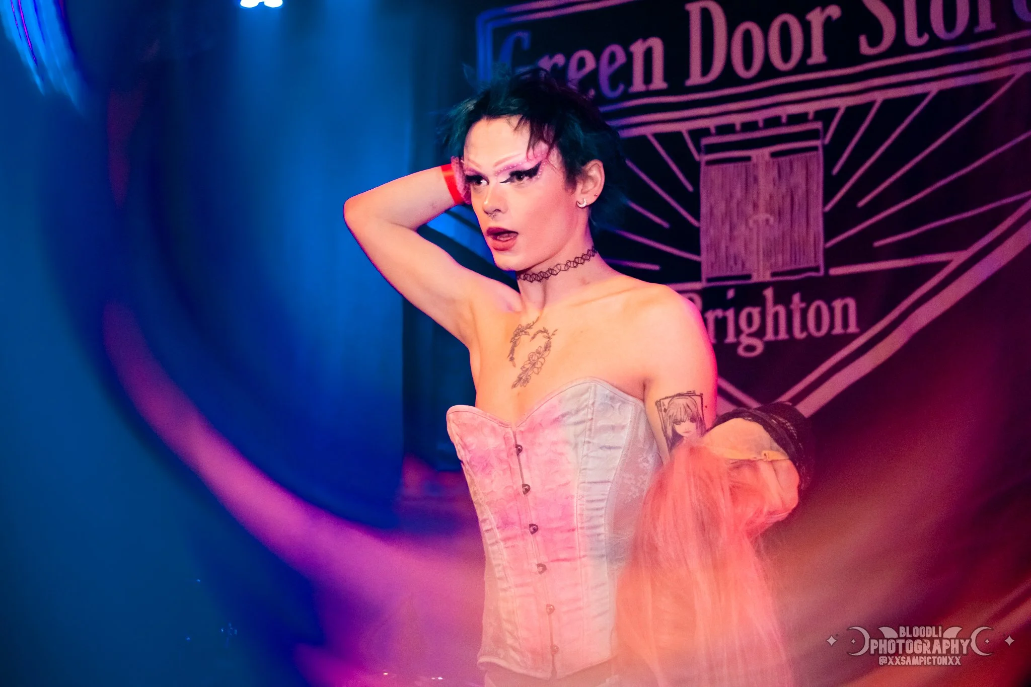 A woman with short black hair and tattoos on her arm, wearing a pink corset and a choker necklace, poses on stage with her hand behind her head, in front of a sign that reads 'Green Door Store' and 'Brighton'.