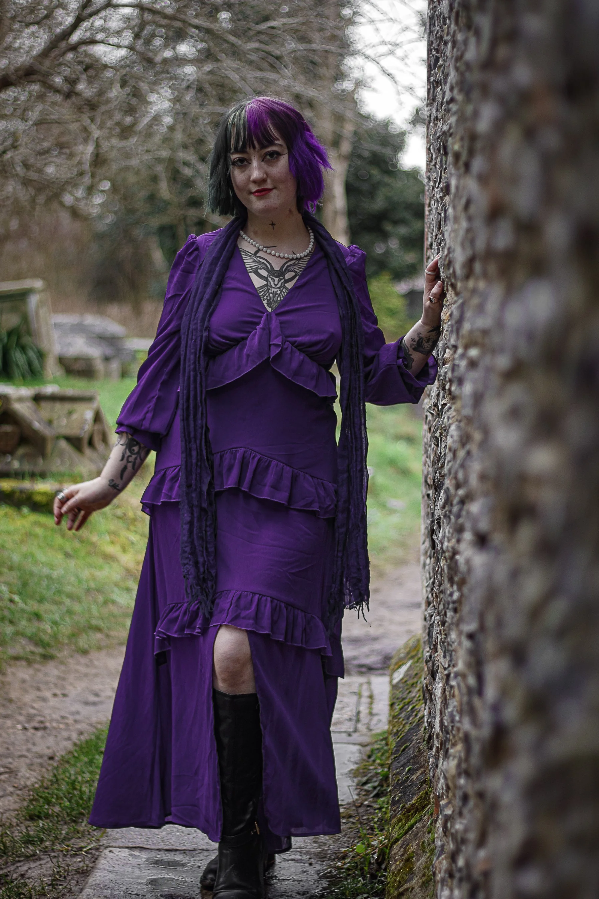 A woman with purple and black hair, dressed in a purple dress with ruffles, standing outdoors next to a stone wall.