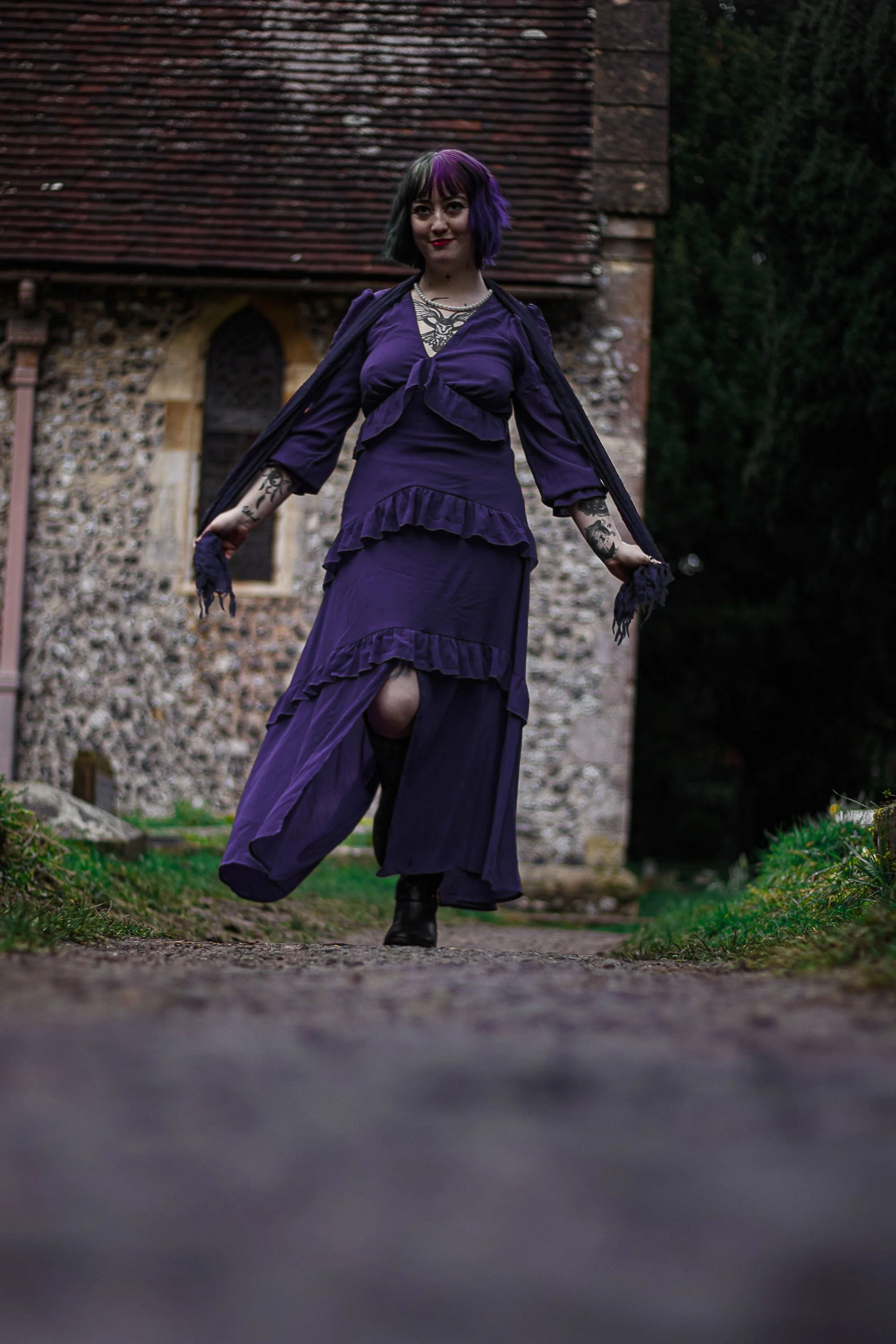 A woman with multicolored hair and tattoos walking towards the camera in a long purple dress outside near a stone building.