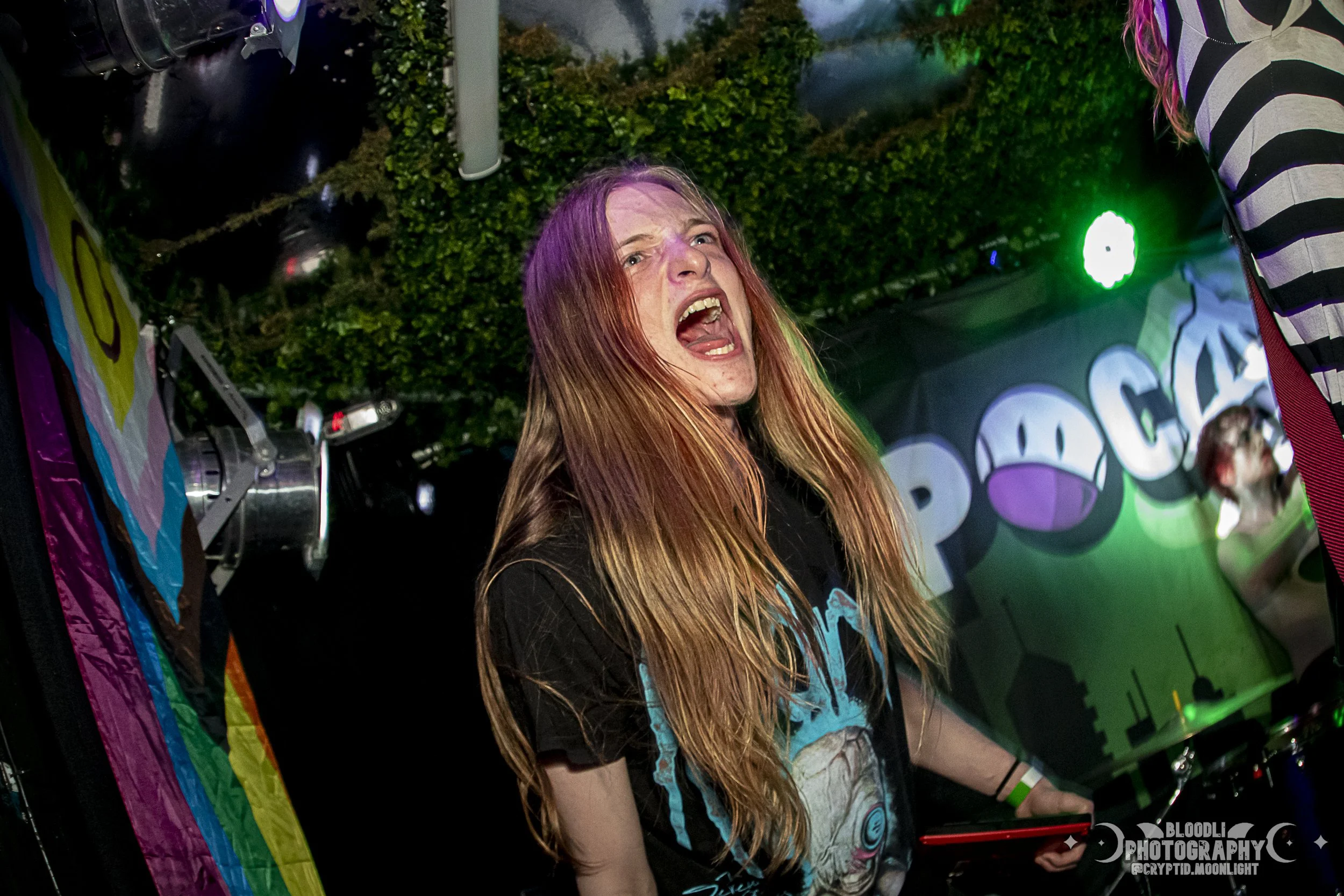 A woman with long, colorful hair singing passionately at a music concert, standing at a DJ booth with a vibrant, colorful background and stage lighting.