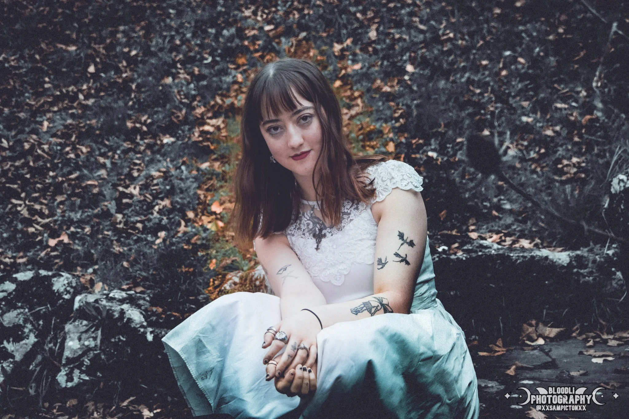 A young woman with dark hair, tattoos, and dark lipstick, dressed in a white lace dress, sitting on a rock surrounded by forest foliage.