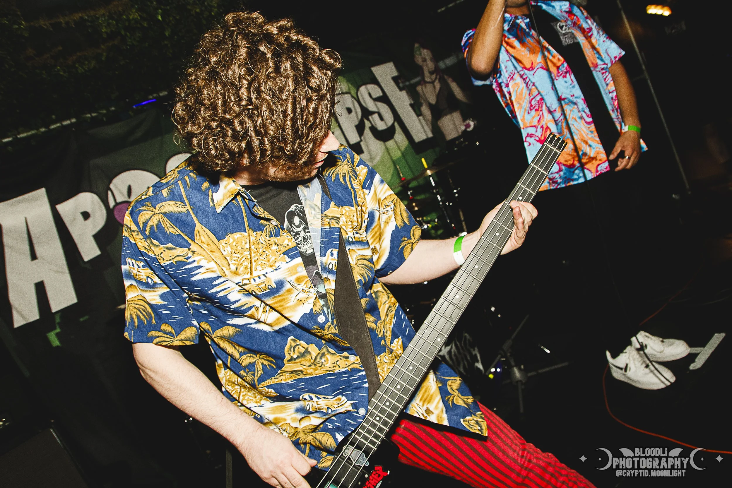 A person with curly brown hair wearing a colorful Hawaiian shirt and red striped pants playing a bass guitar at a concert or music event.