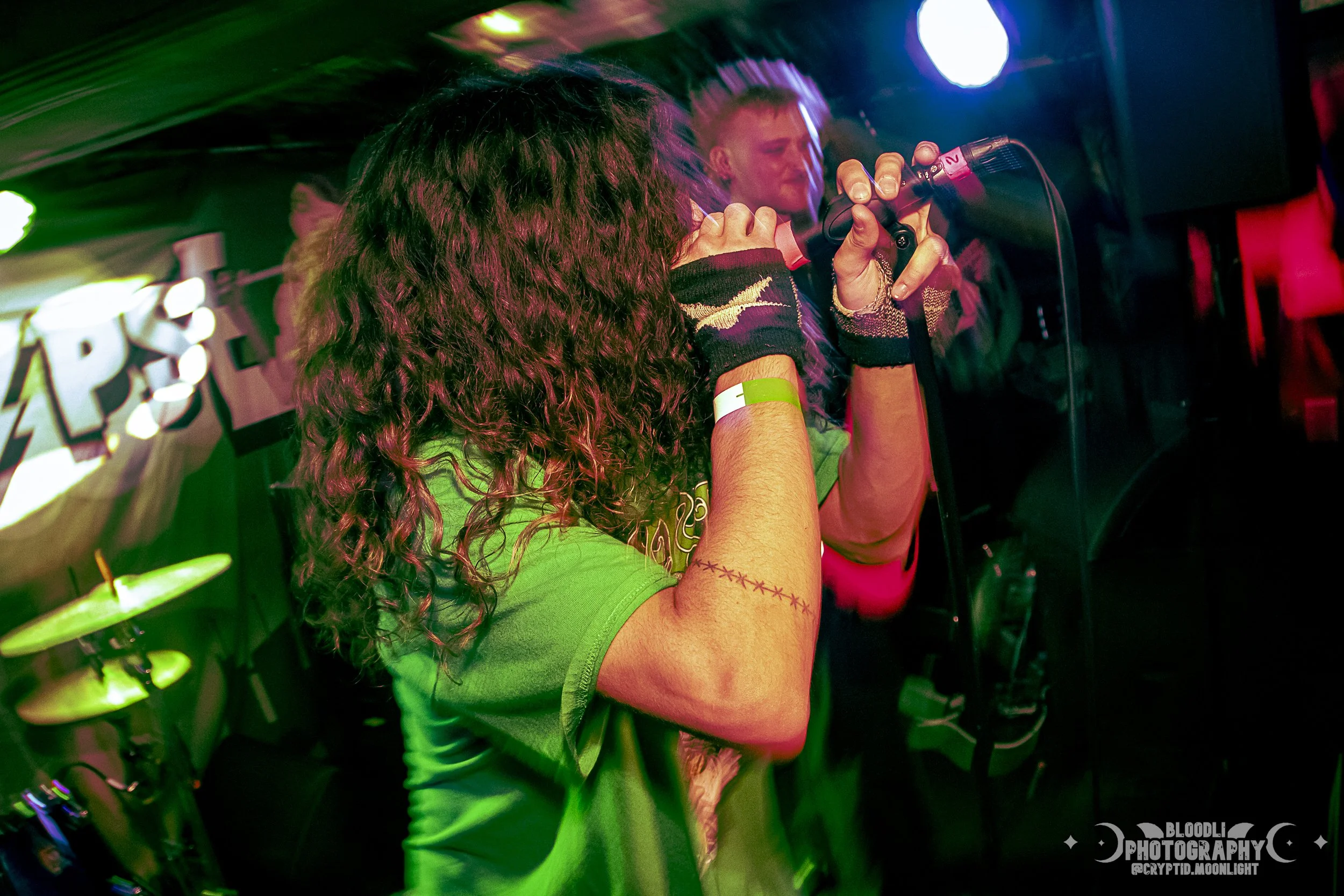 Person with long curly hair singing into a microphone at a concert or karaoke event, with colorful stage lighting and a banner in the background.