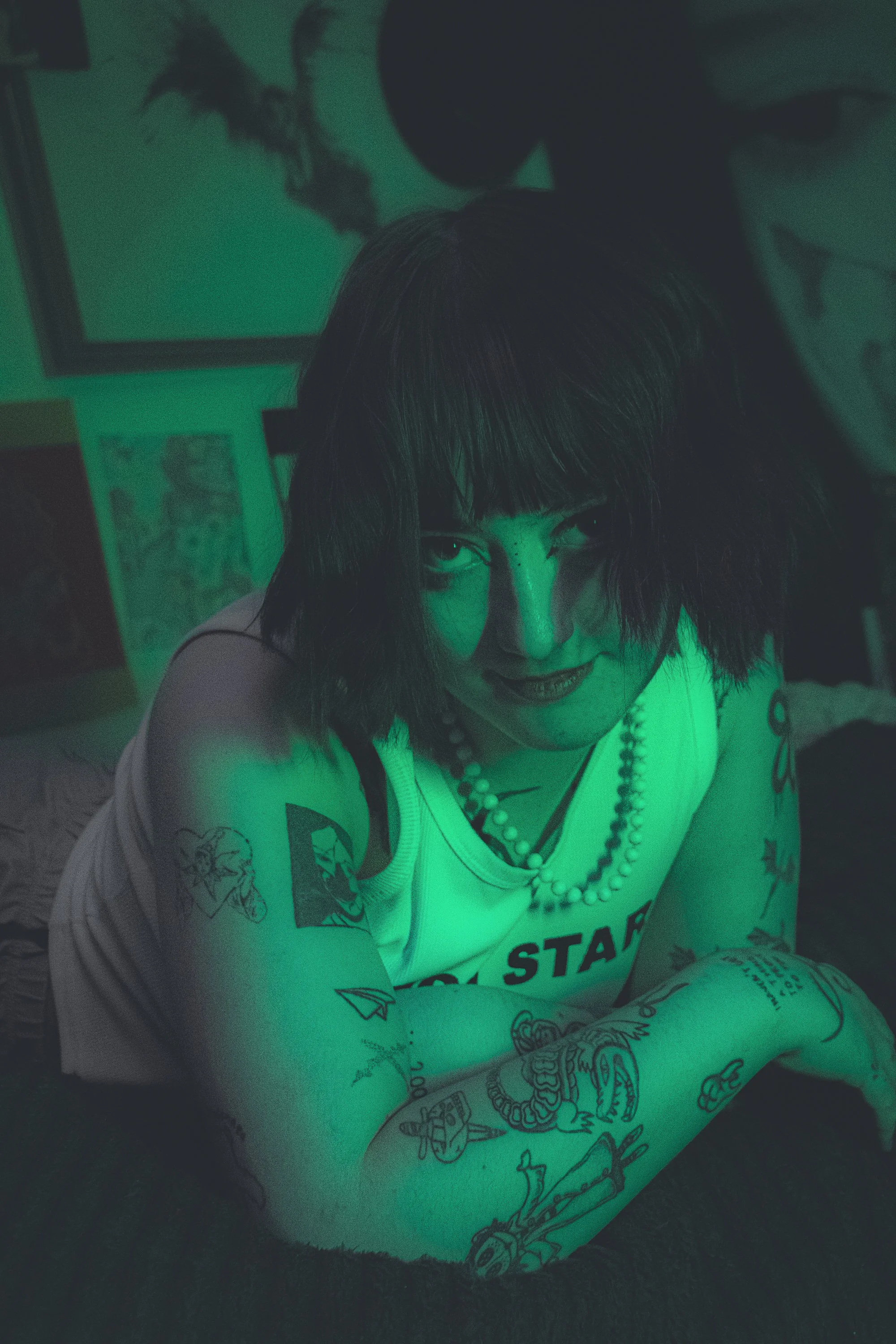 A person with tattoos and dark hair, leaning forward with a serious expression, illuminated by green lighting in an art-filled room.