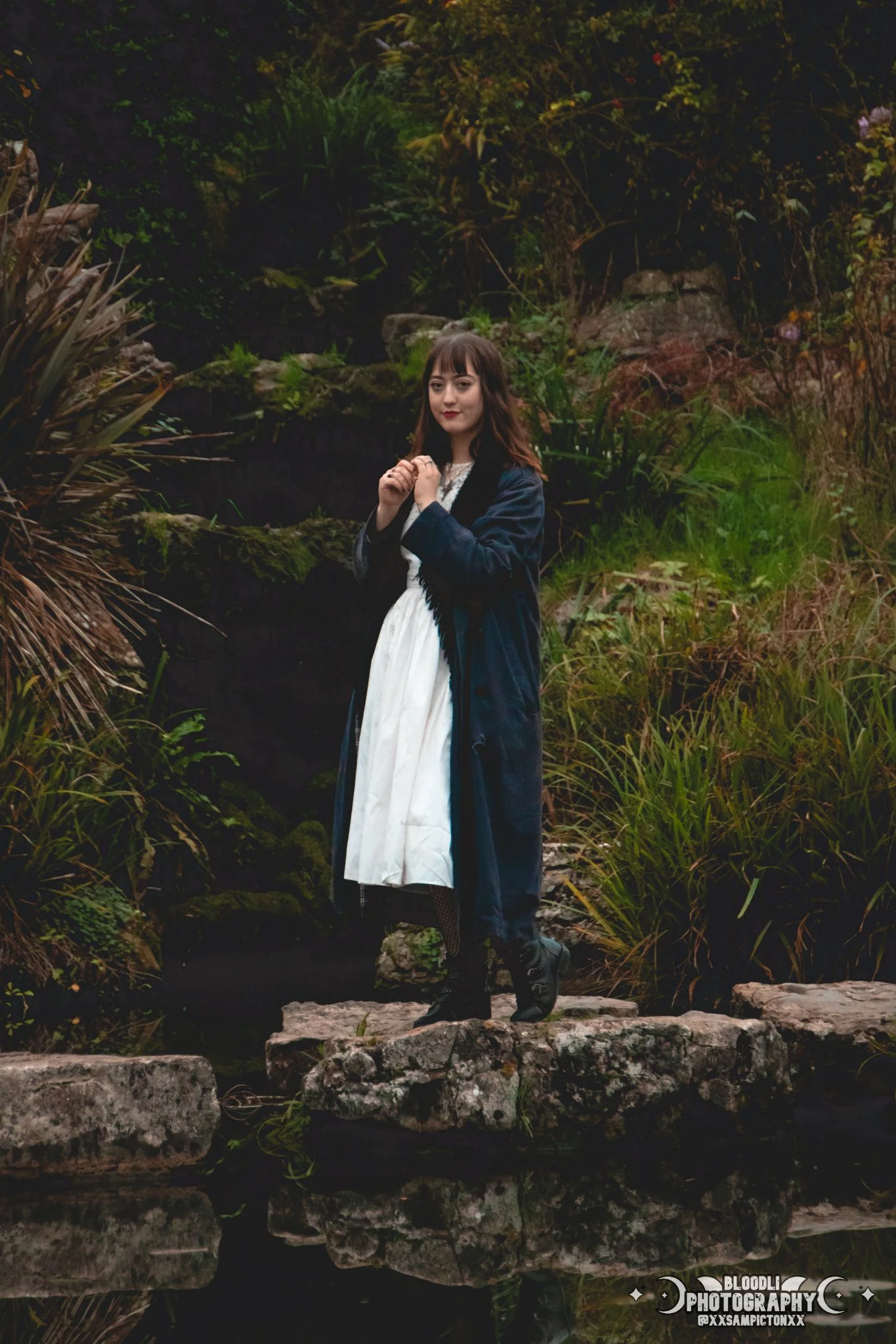 A woman standing on rocks by a pond in a lush, green garden. She is wearing a white dress and a long dark coat, with her hands clasped near her chest and looking at the camera.
