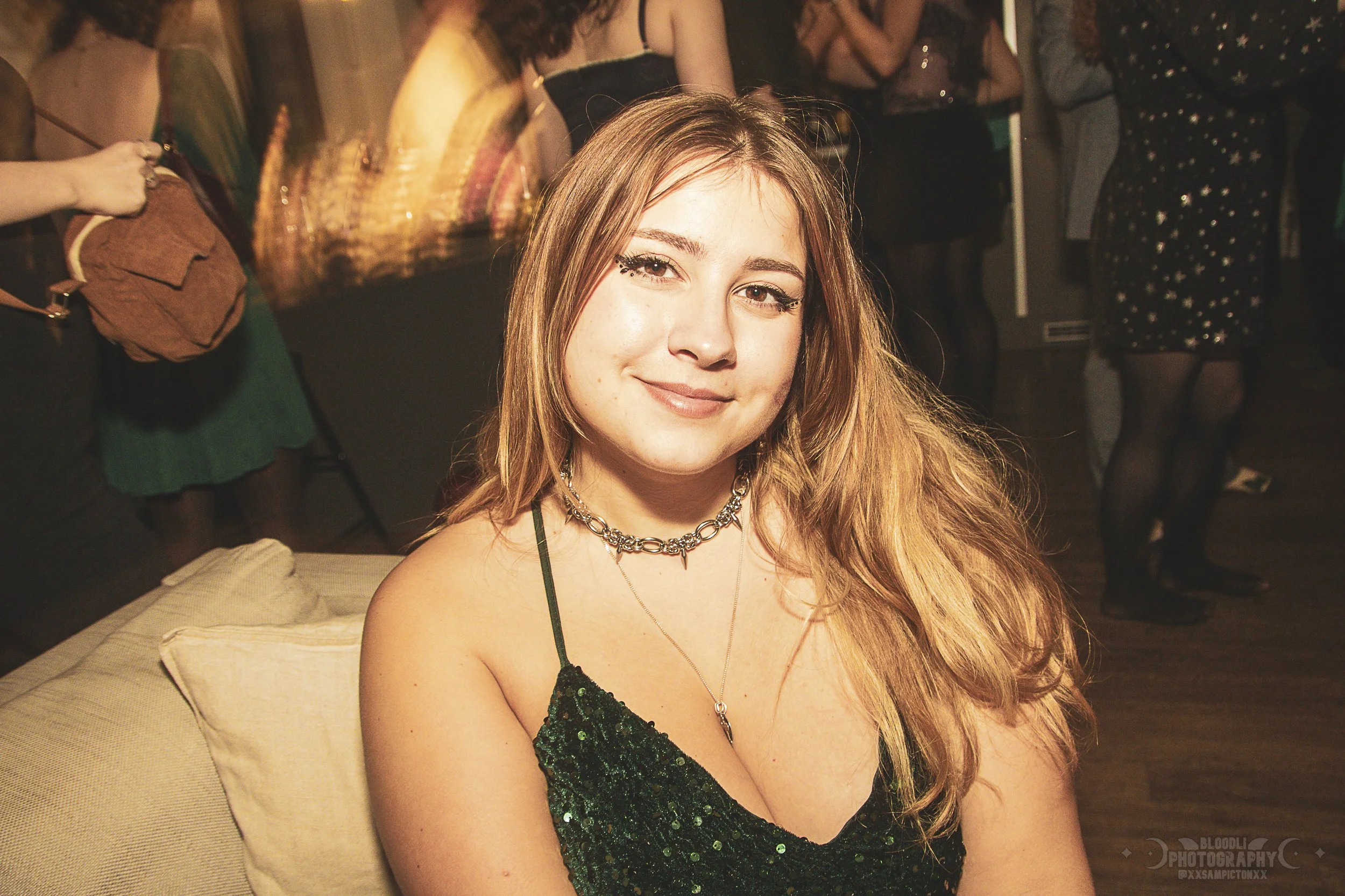 Young woman with blonde hair sitting on a beige couch, smiling at the camera in a dimly lit party or club setting with other people in the background.