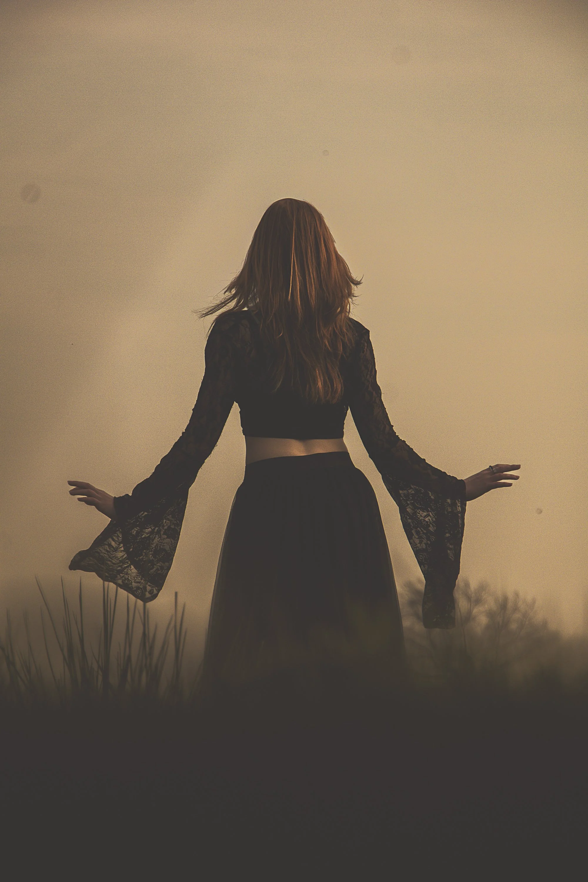 A woman with long, wavy hair standing outdoors during sunset or dusk, wearing a black dress with lace sleeves and a midriff-baring top.