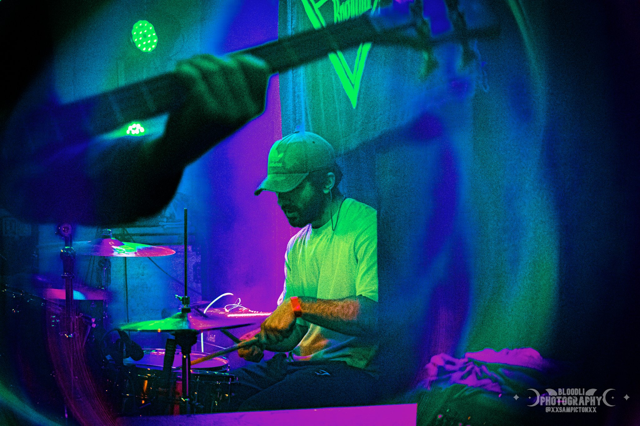 A person playing drums in a room with neon lights, wearing a baseball cap, white shirt, and a smartwatch.
