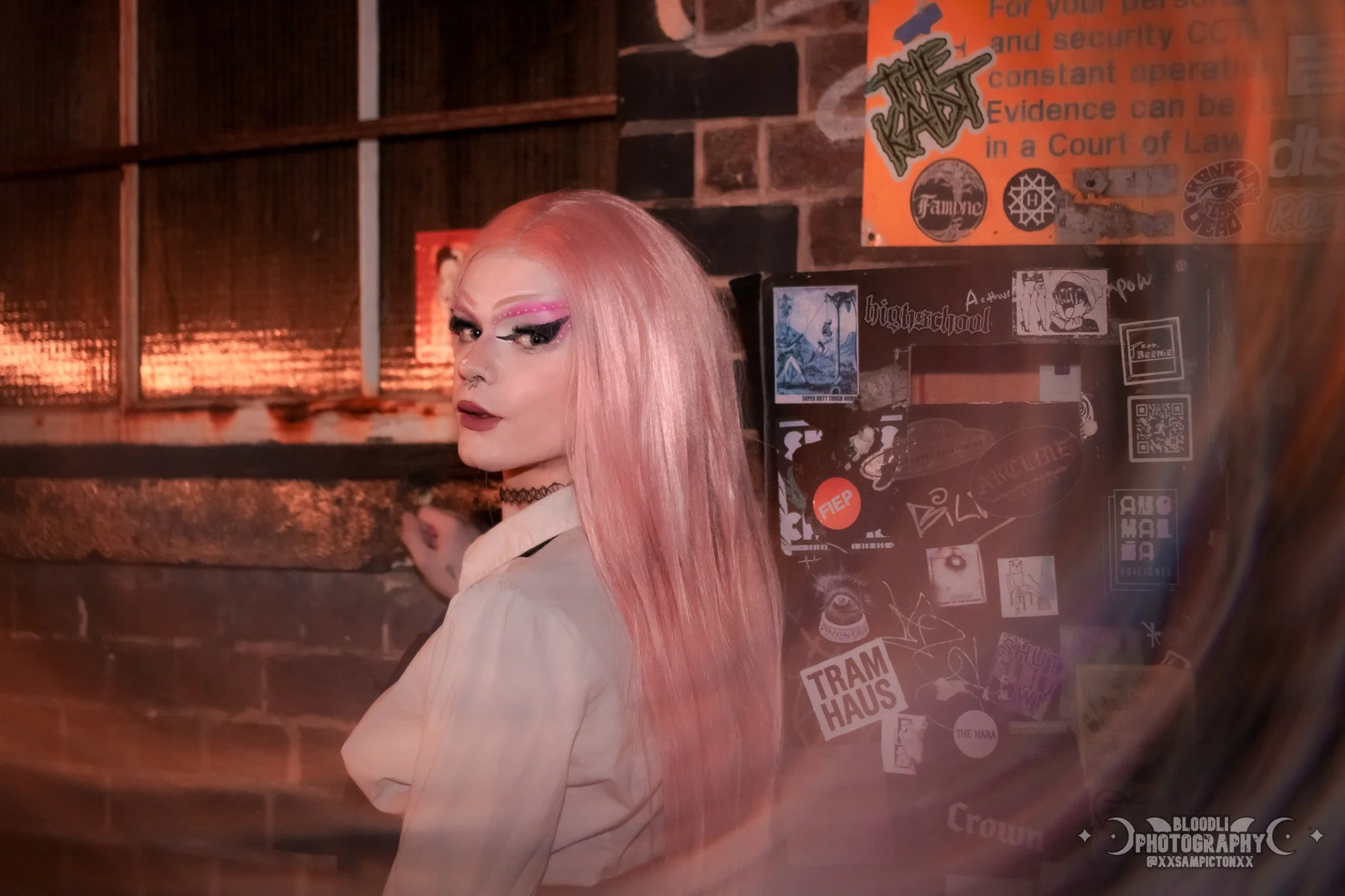 A person with long pink hair and bold makeup, including pink and black eyeshadow, poses in front of a brick wall decorated with stickers and posters in a dimly lit room.