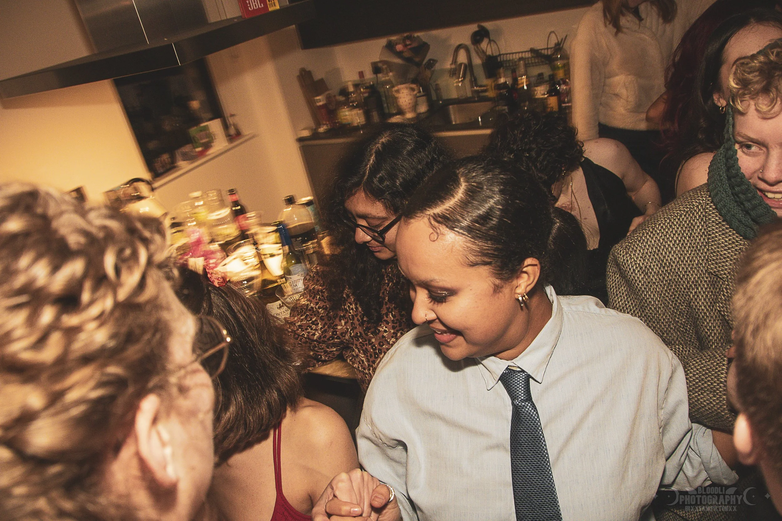 People socializing at a party in a kitchen, with drinks and food on the counter, and a warm, lively atmosphere.