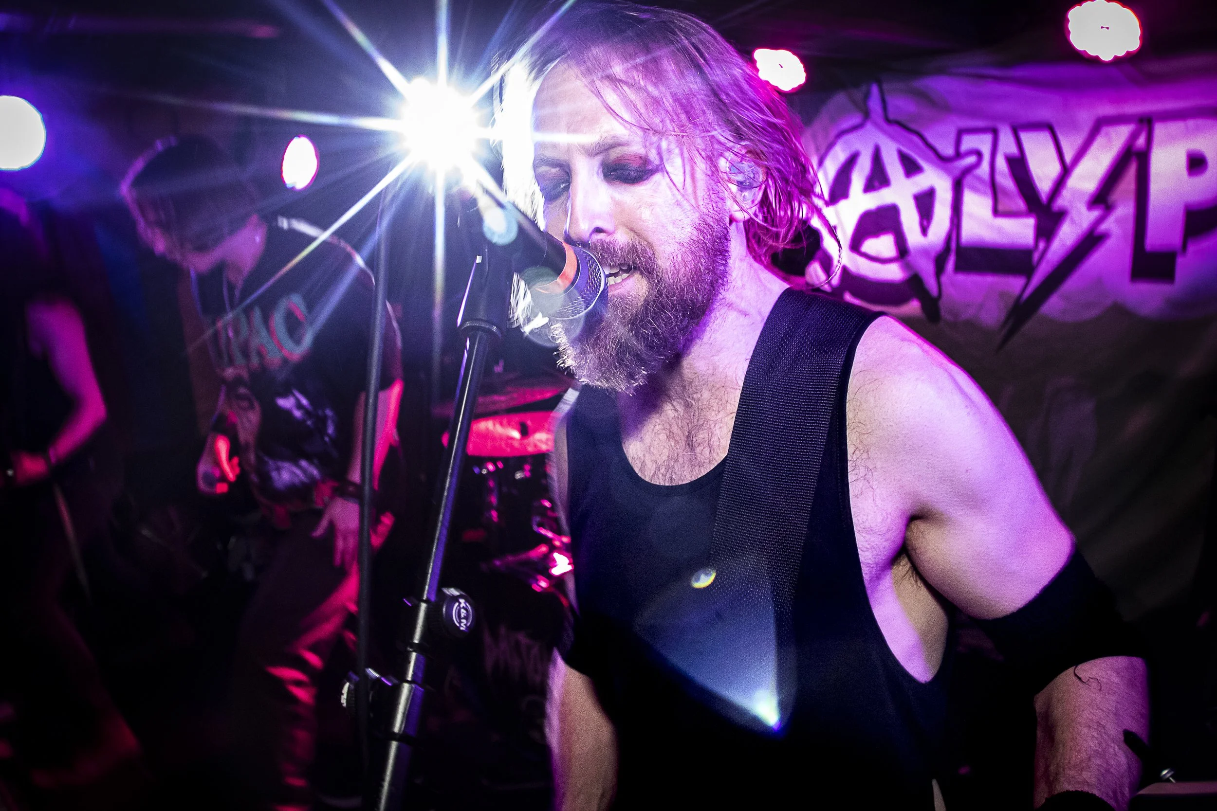 A male vocalist with a beard and long hair singing into a microphone on stage, illuminated by purple and pink lights, with a band member in the background and a banner reading 'GATE' in stylized letters.