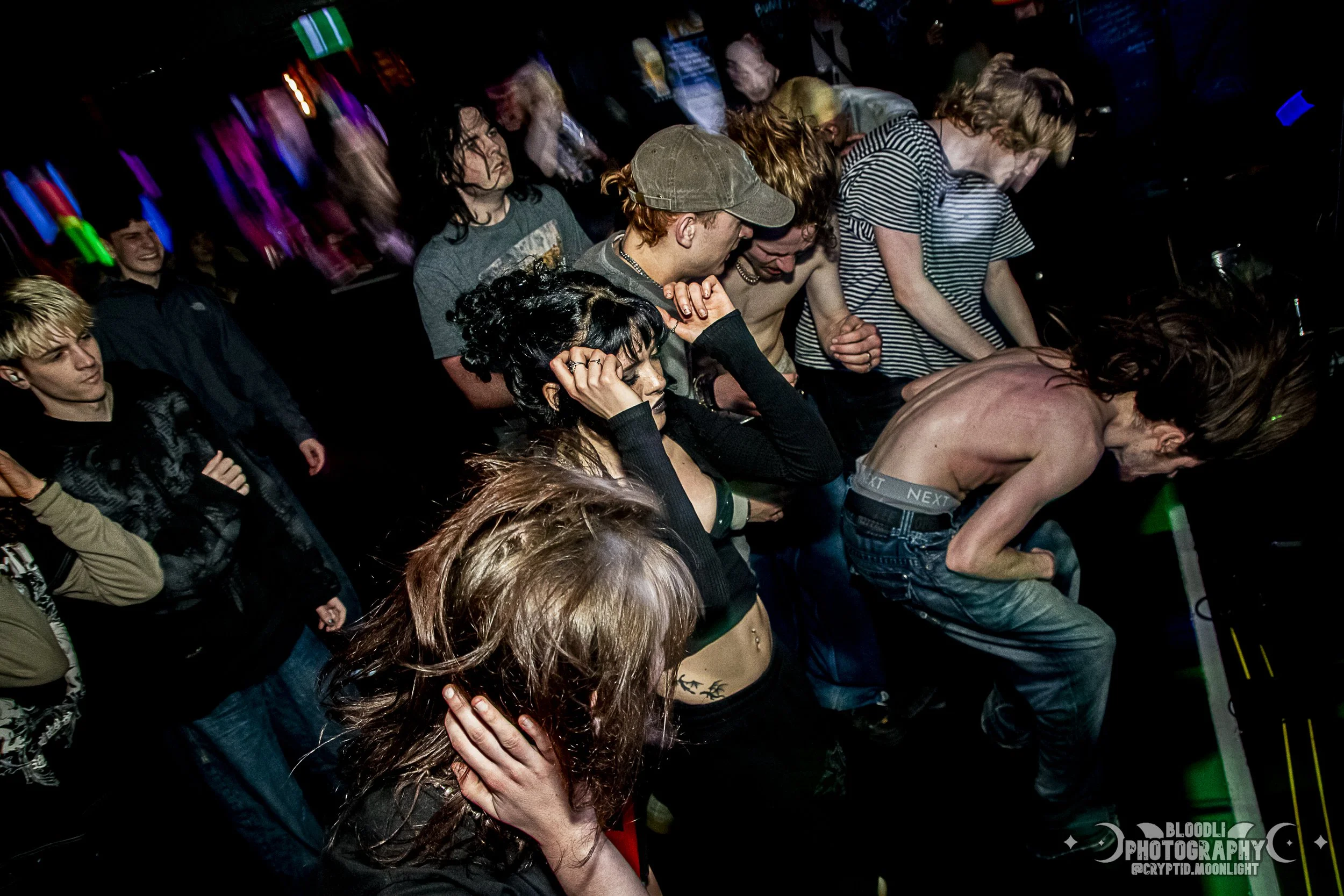 Group of young people dancing and enjoying music at a concert or nightclub with colorful lighting.