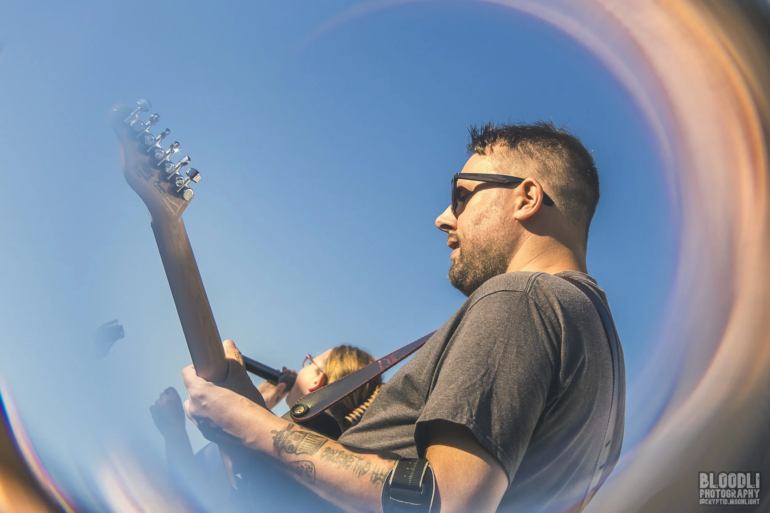 A man with tattoos and sunglasses playing an electric guitar during a sunny outdoor event, with a rainbow-like reflection in the foreground.