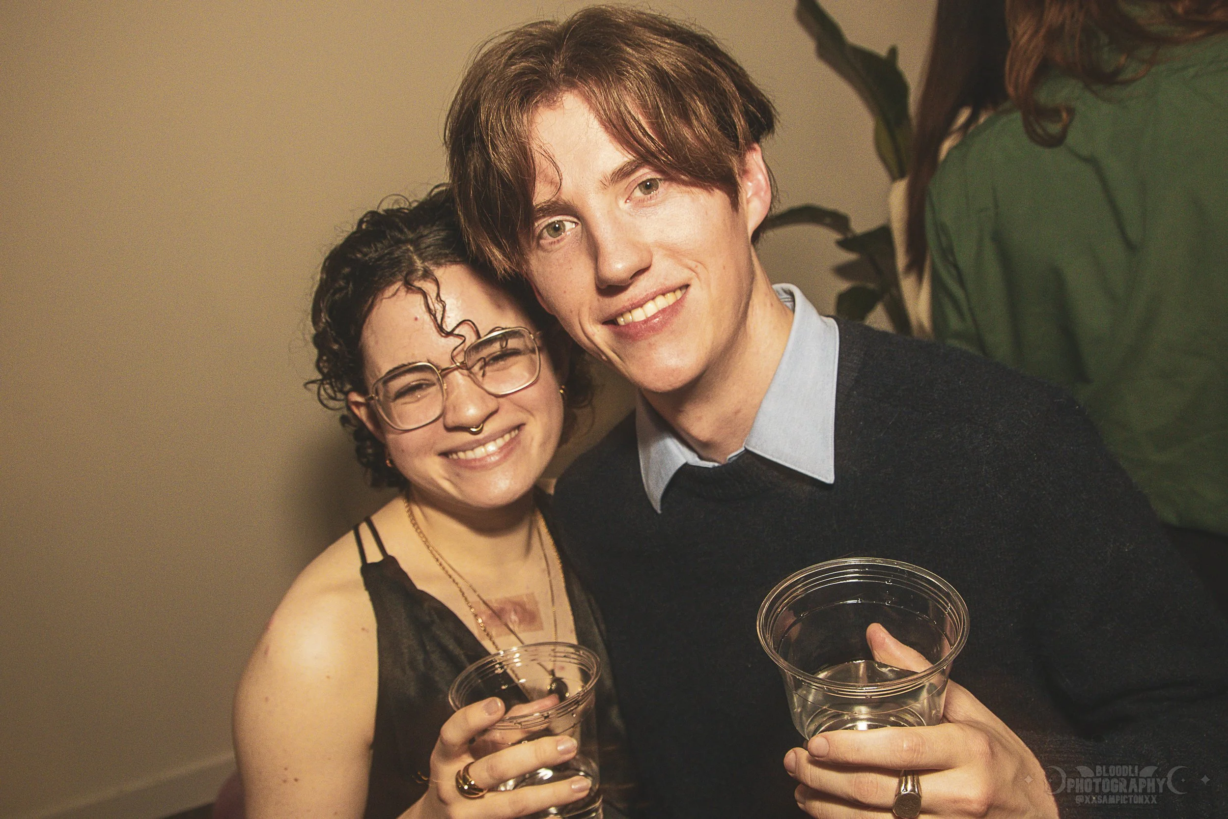 Two young adults smiling and holding drinks at a social gathering. The woman has short, curly hair, glasses, and piercings, and the man has medium-length brown hair.