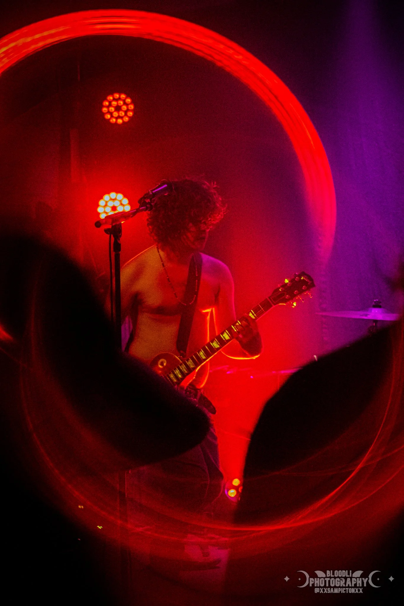 A shirtless male guitarist with curly hair playing an electric guitar on stage, illuminated by red and purple stage lights, with a circular light pattern behind him, and a cymbal partially visible on the right.