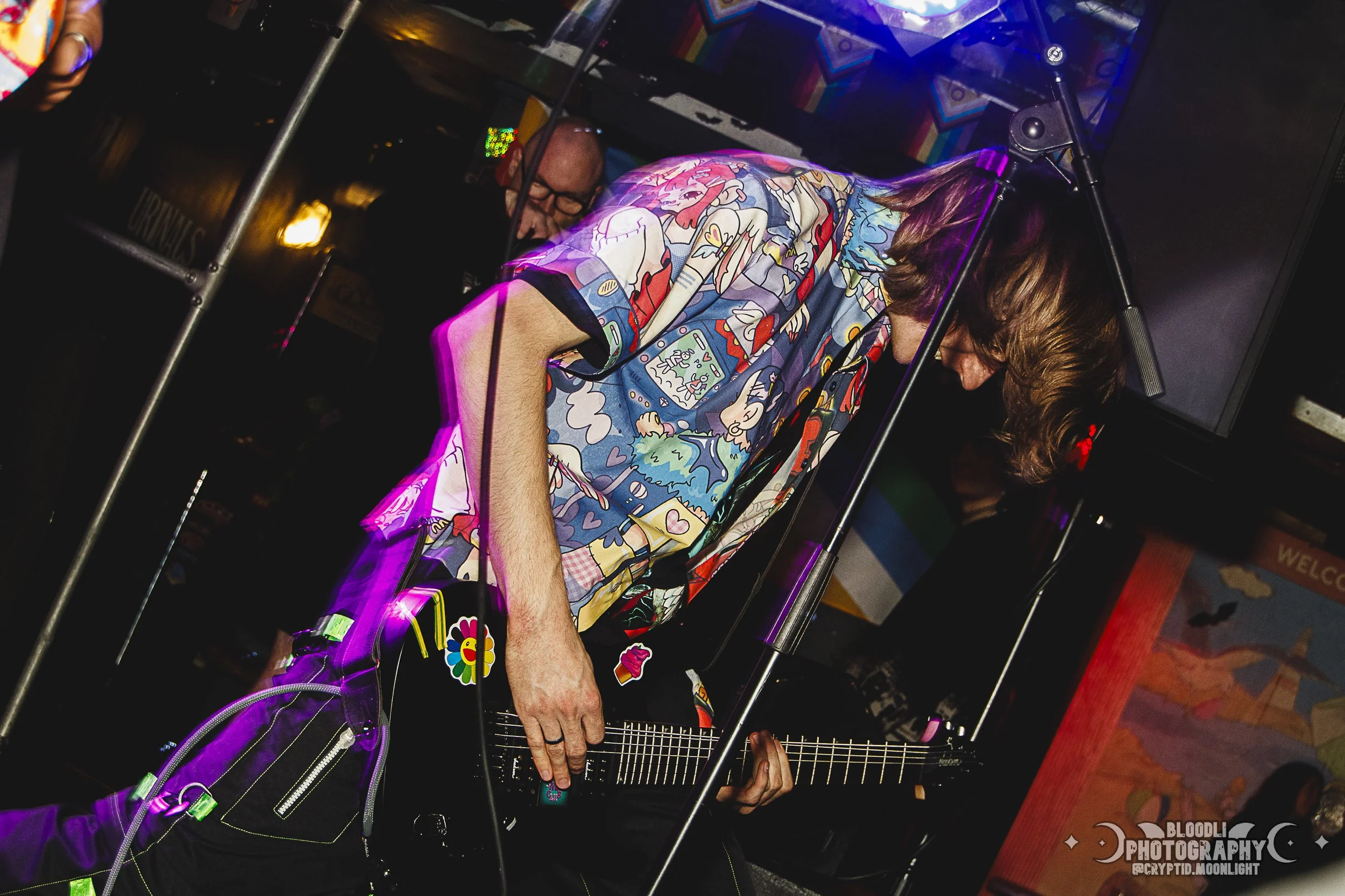 A person with long brown hair wearing a colorful, patterned shirt playing an electric guitar on stage, with a man and a woman visible in the background.